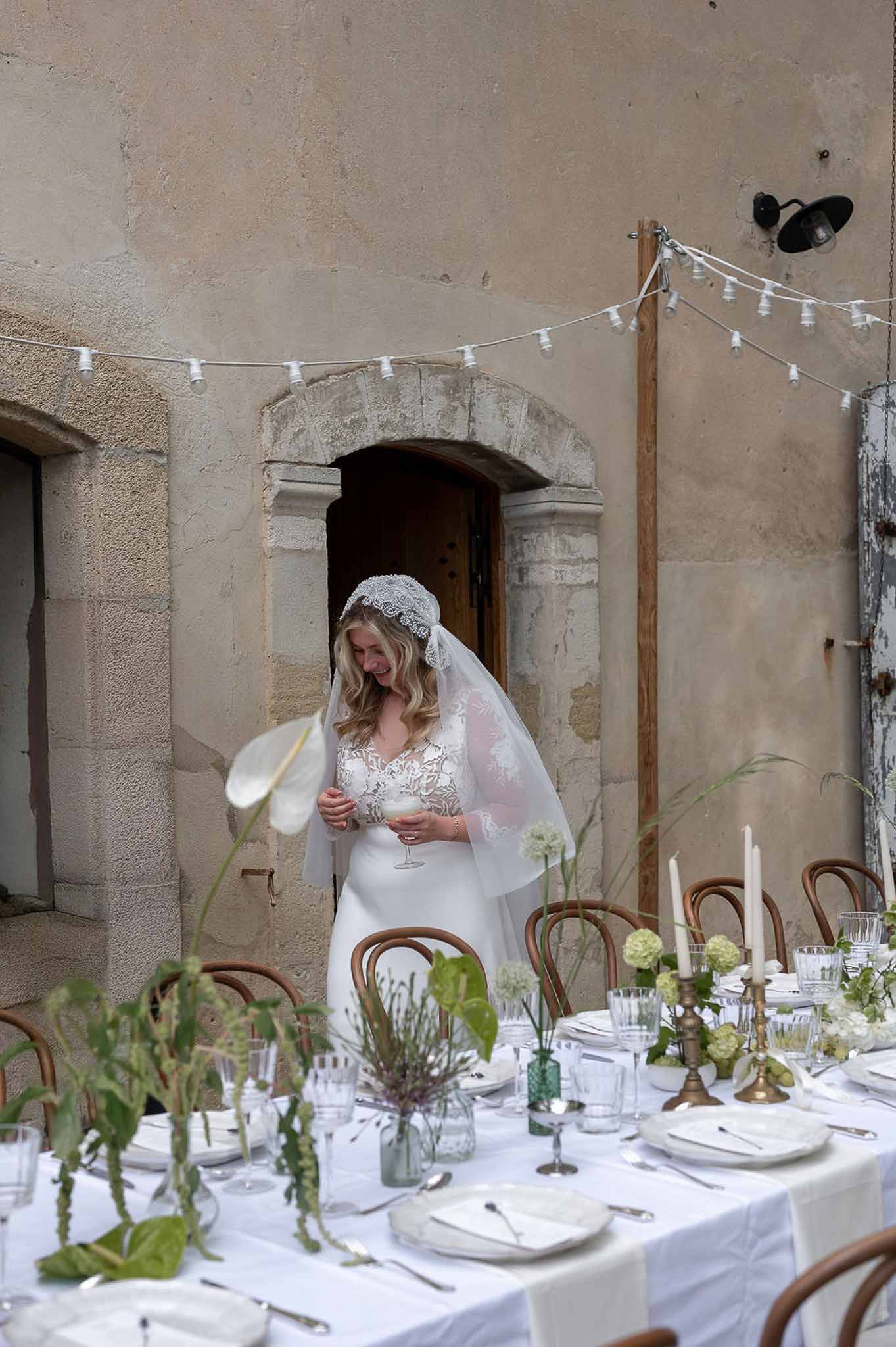 Bride in lace gown and Juliet-cap veil beside table with anthurium and hydrangea bud vases in courtyard
