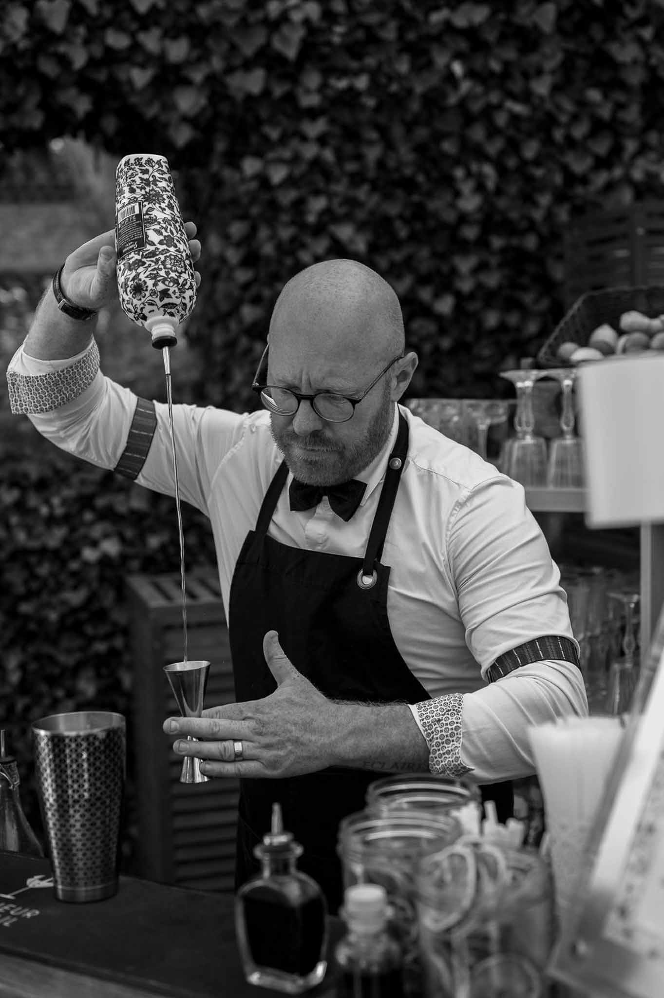 Bartender in white shirt and black bow tie pouring cocktail from jigger at outdoor bar in black and white