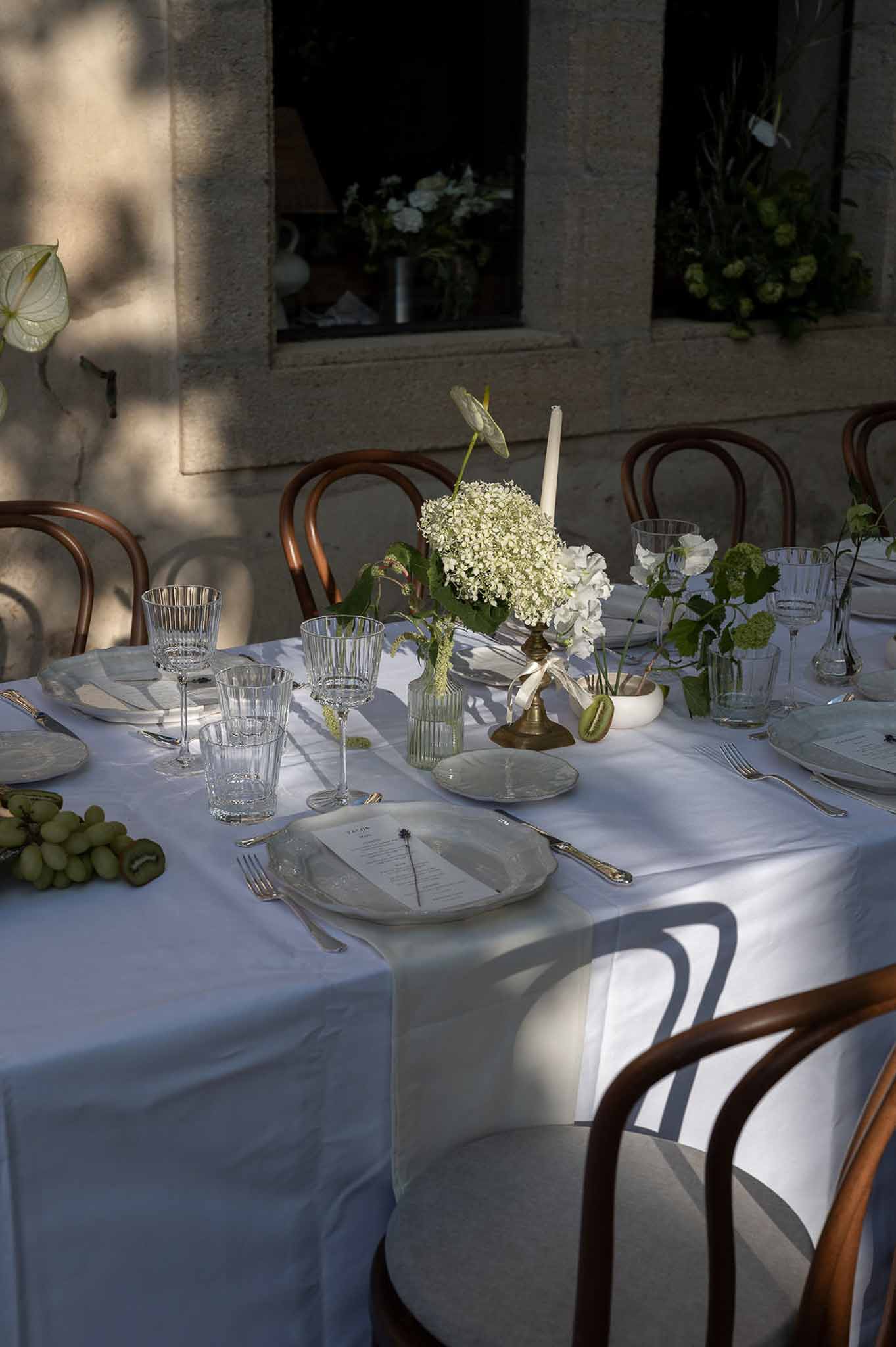 Round table with hydrangea brass candlestick, grapes, kiwi, and bud vases in dappled sunlight