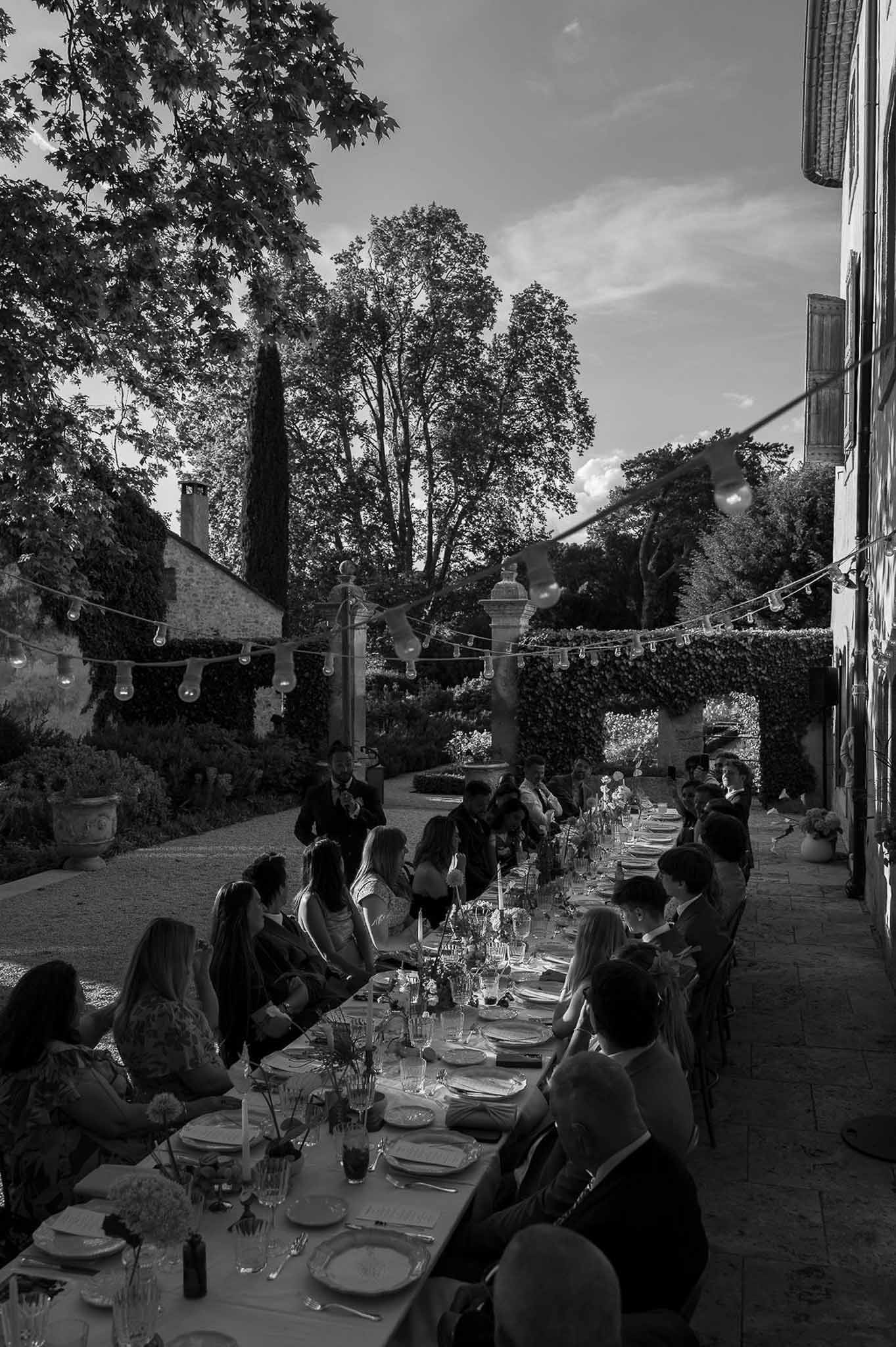 Black and white long reception table with Edison lights and ivy-covered courtyard wall at evening dinner