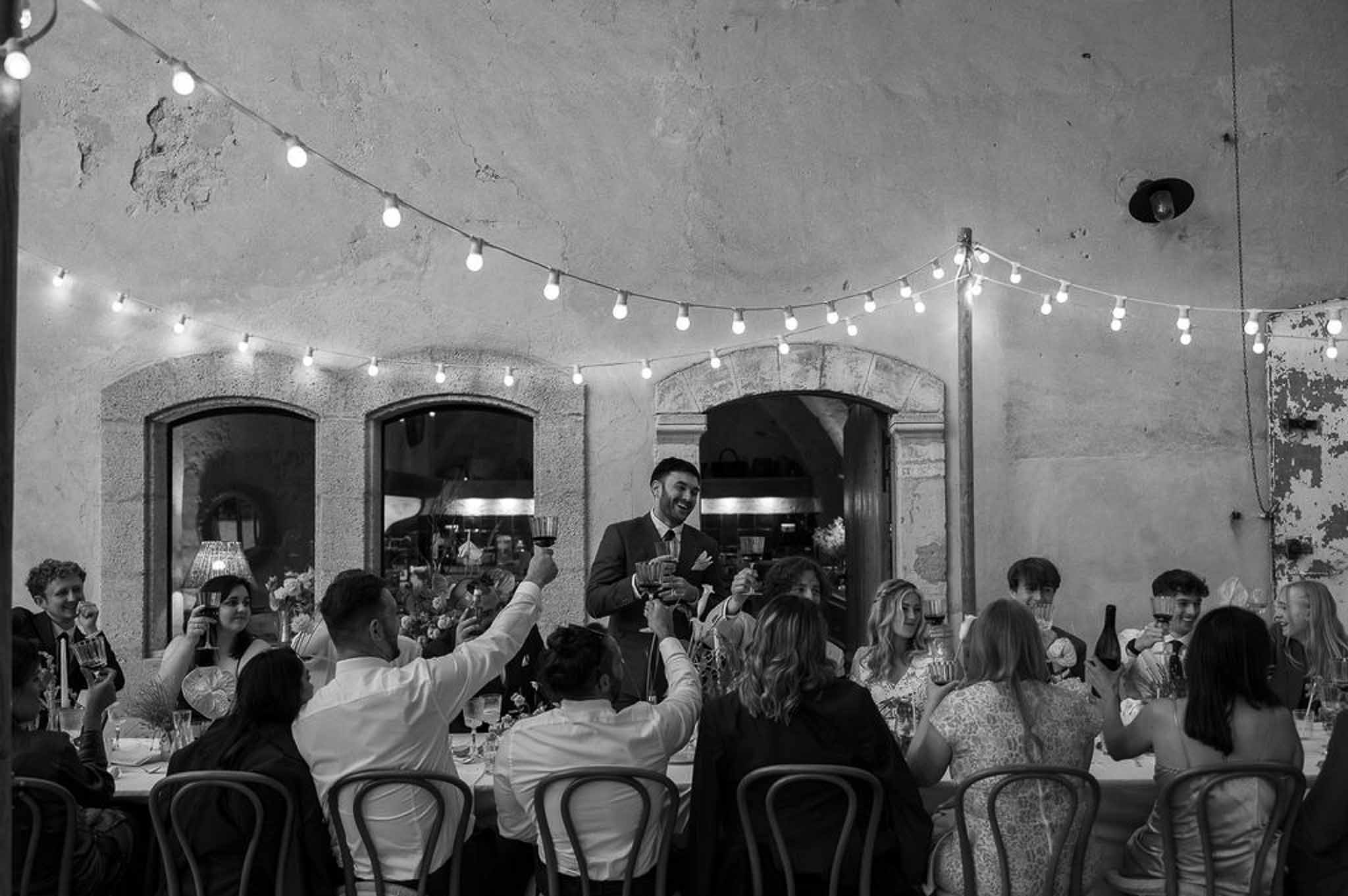 Black-and-white wide shot of guests raising glasses during a toast in a stone-walled venue with fairy lights