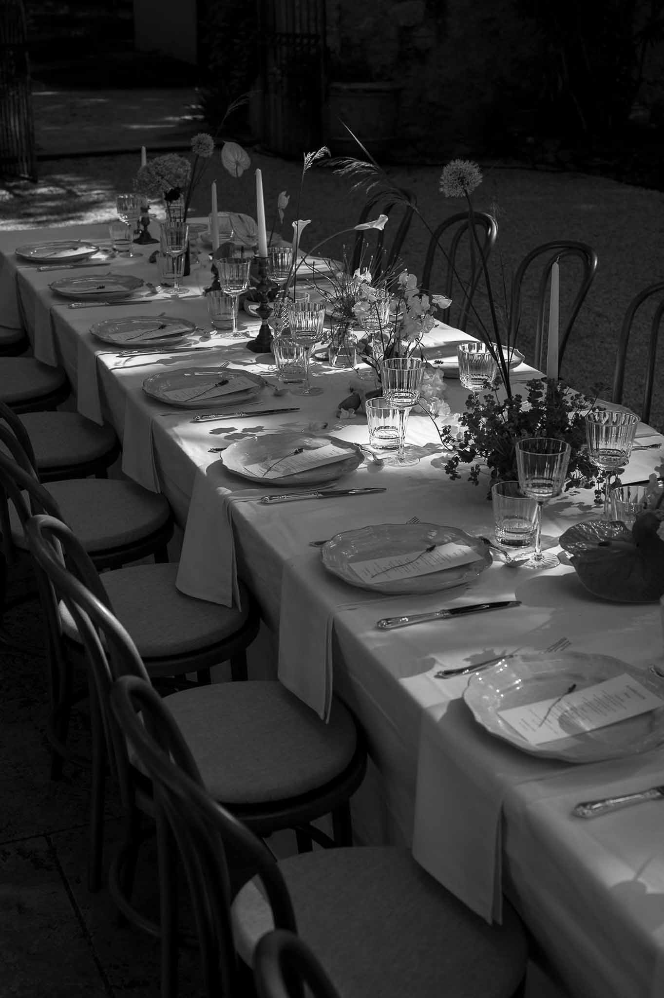 Black-and-white photo of a long outdoor reception table with taper candles, allium, calla lilies, and crystal glassware