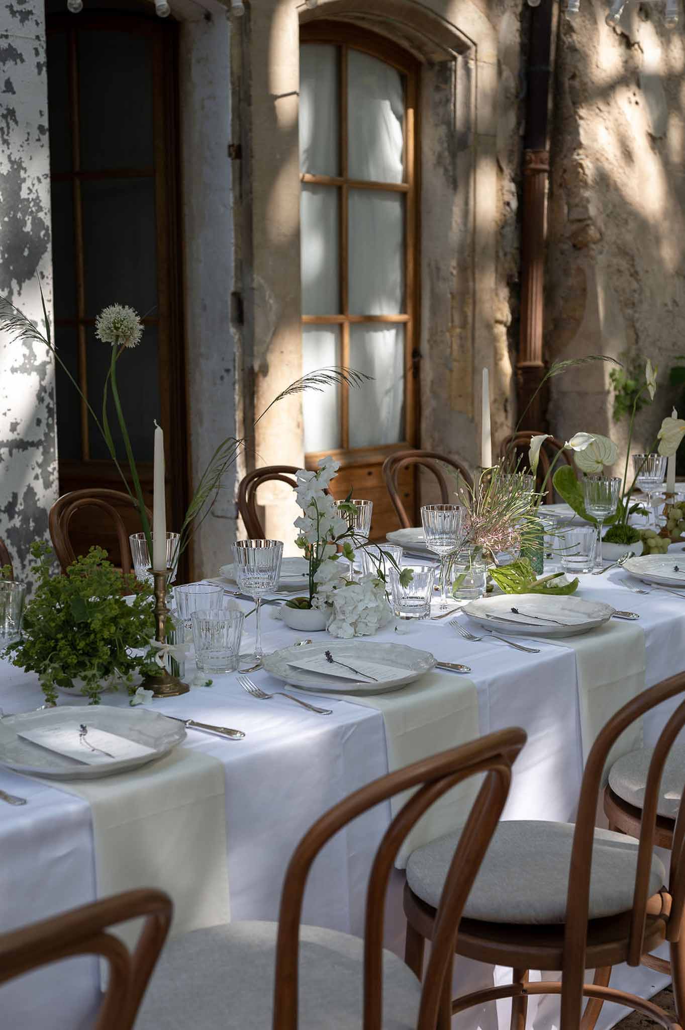 Outdoor reception table with white hydrangeas, orchids, and green foliage centerpiece, crystal glasses, and brass candlest...