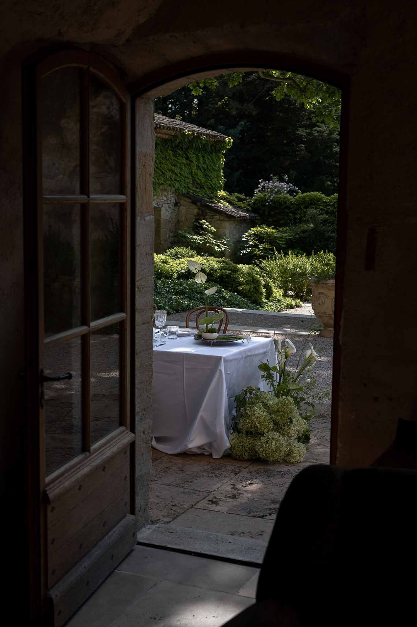 Reception table with white anthurium and calla lily arrangements framed through arched stone doorway at French chateau