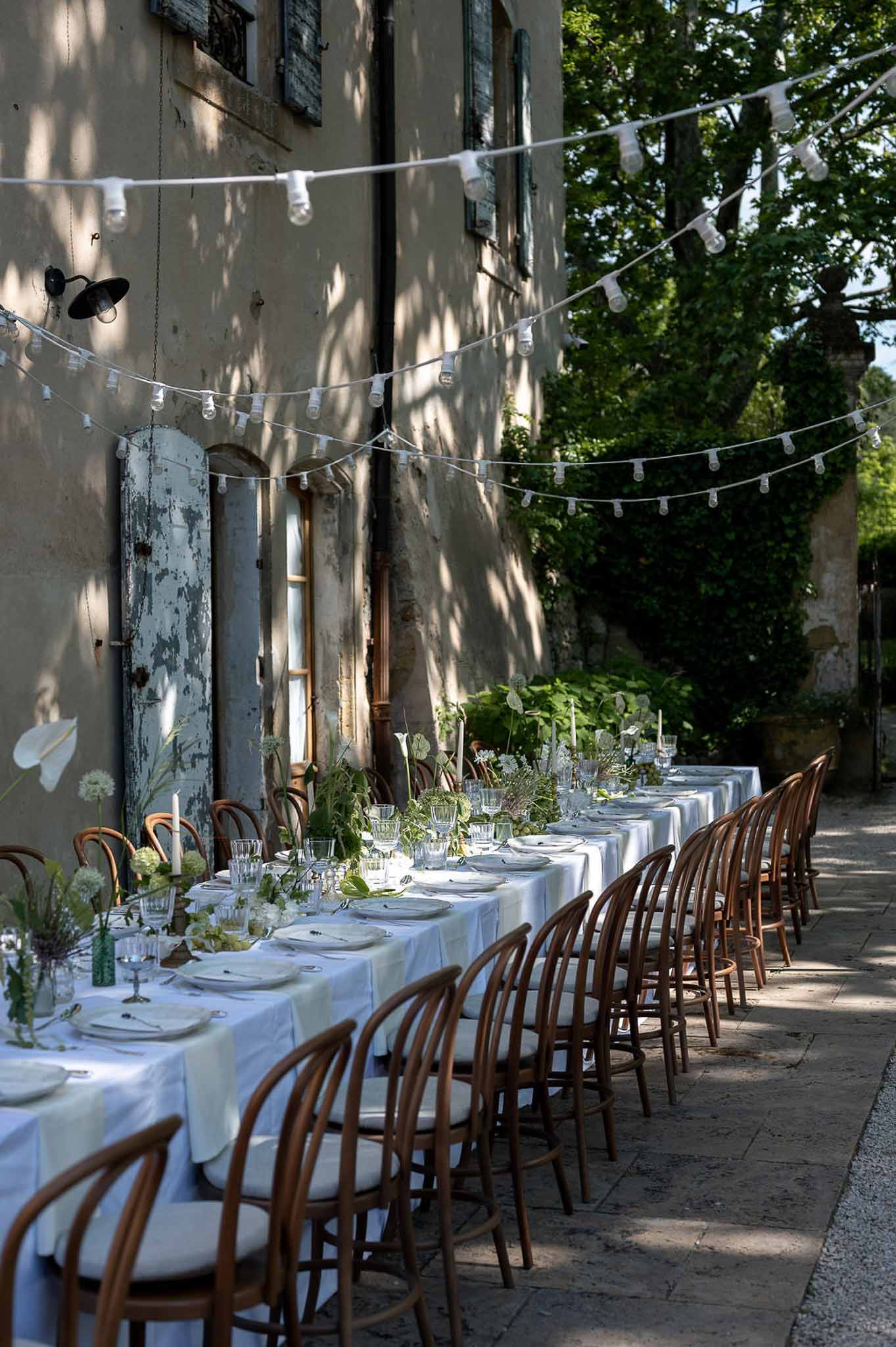 Long outdoor banquet table with white linen, green and white floral runner, taper candles, and festoon lights