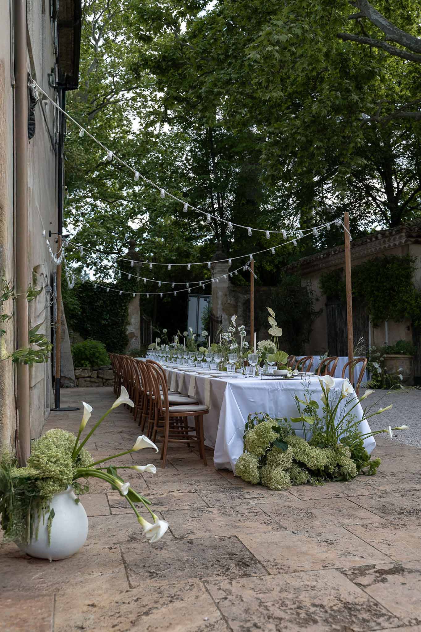 Long reception table with white calla lilies and green hydrangeas under string lights in French stone courtyard