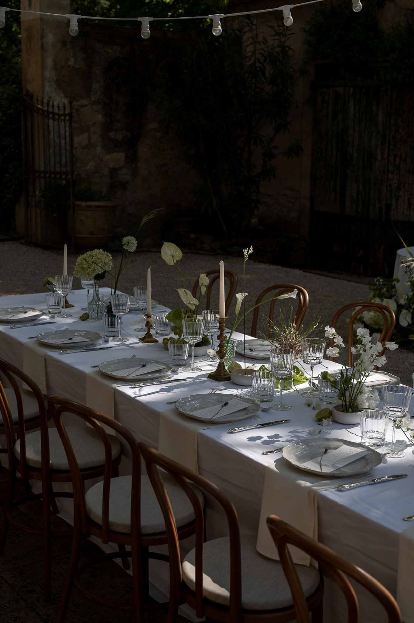 Reception table with white hydrangeas, anthurium, taper candles in brass holders, and festoon lights overhead