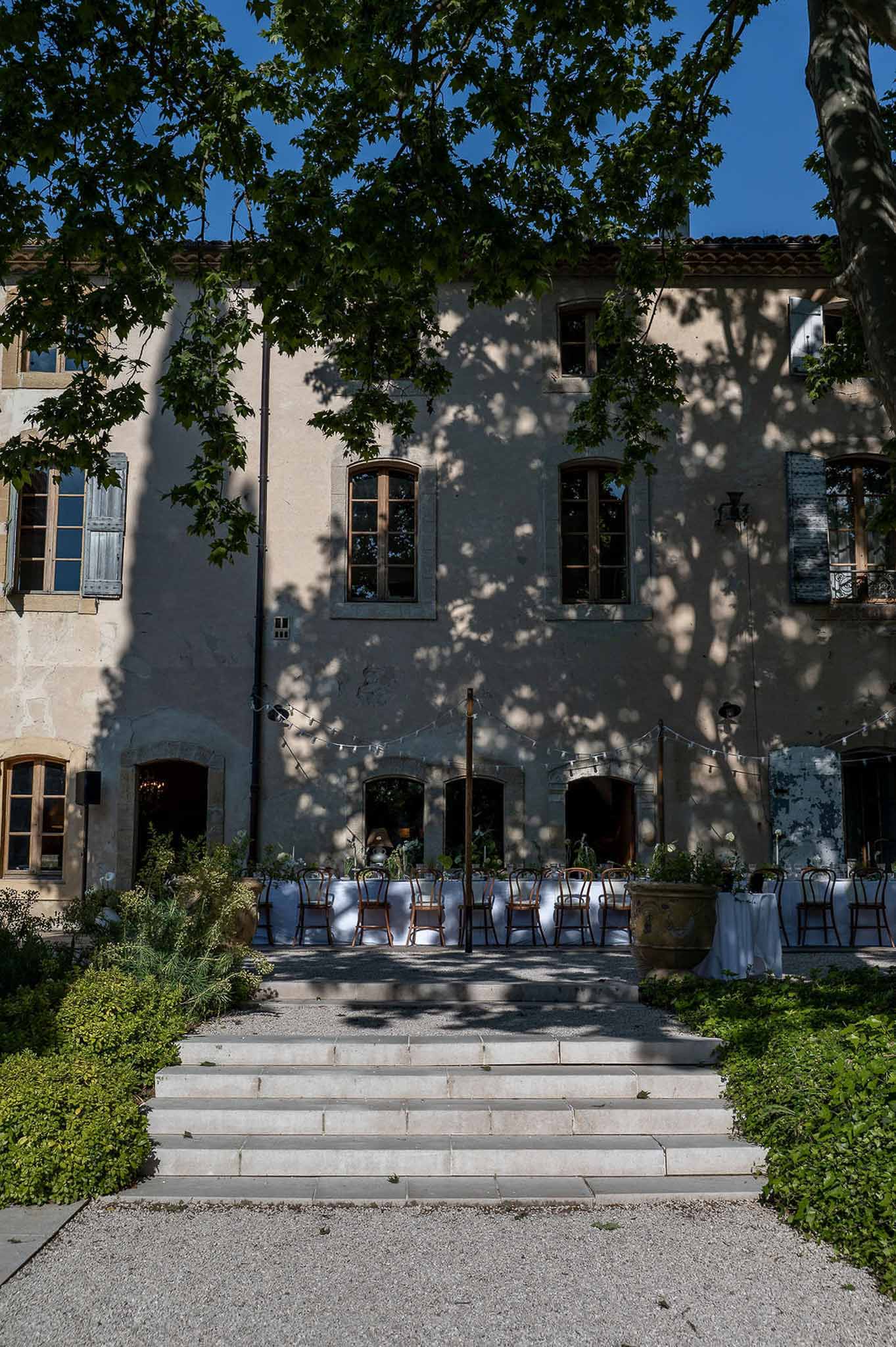 Long reception tables with bentwood chairs on manor terrace with string lights and dappled tree shadow