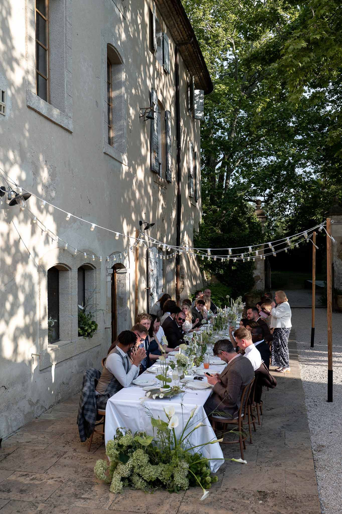 Twenty-five guests at banquet table with calla lily and viburnum runner festoon lights against stone manor