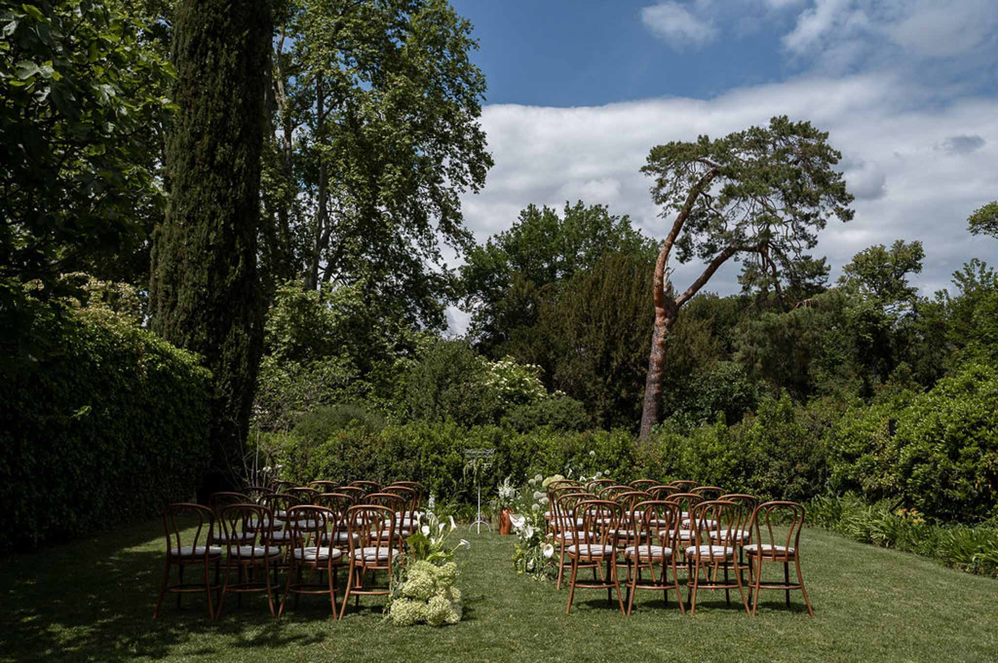 Empty outdoor ceremony setup with chestnut bentwood chairs and white floral arrangements on manicured garden lawn