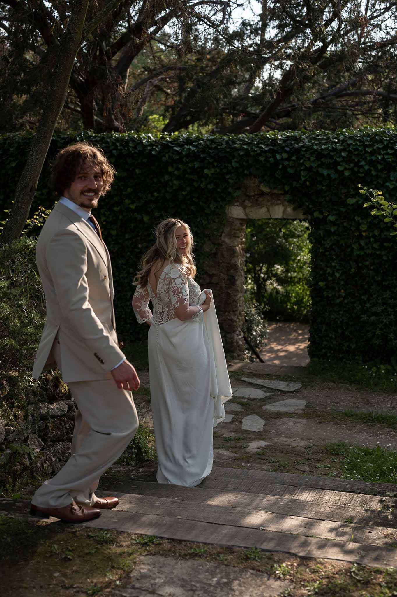 Couple glancing back on garden path bride in lace-bodice gown with open back groom in sand suit under ivy arch