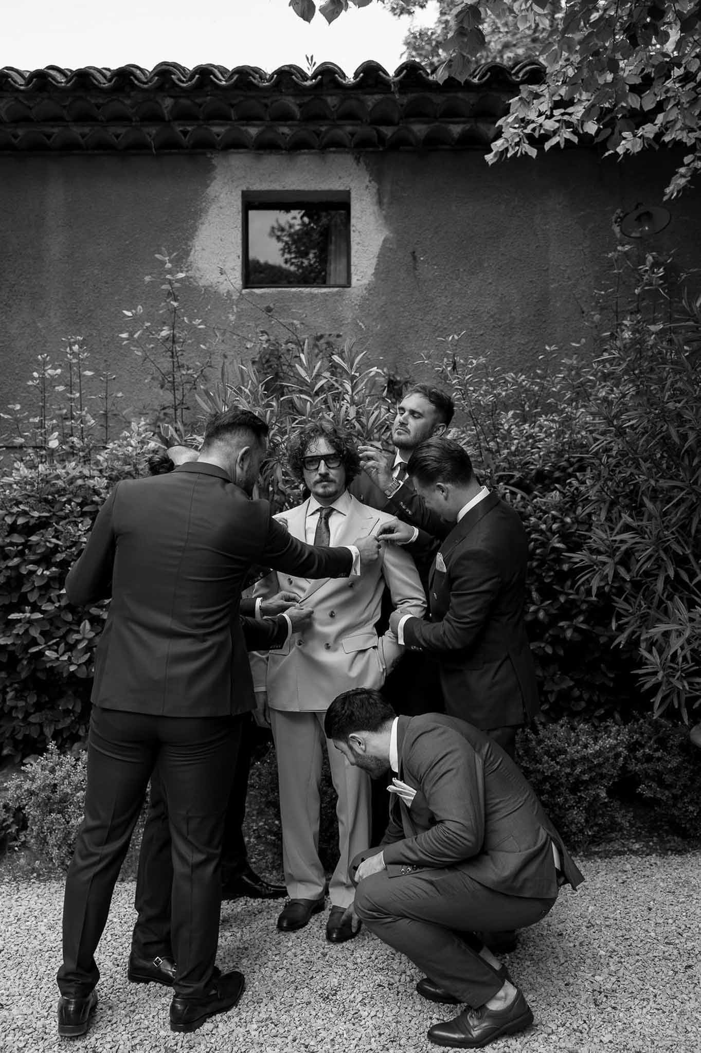 Black and white of four groomsmen adjusting groom's suit simultaneously on gravel path before stone building
