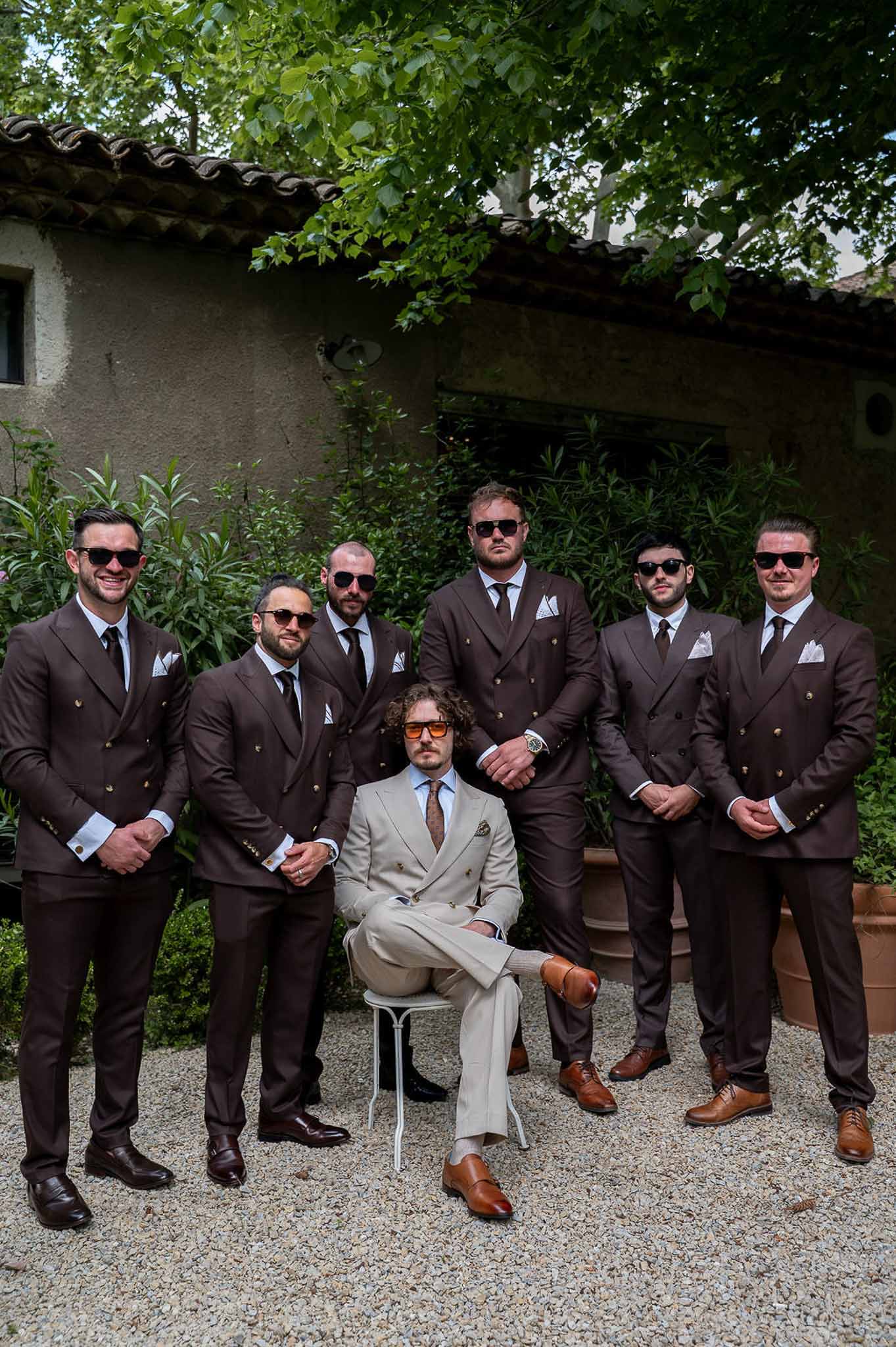 Groom in beige suit seated with six groomsmen in brown double-breasted suits posing on a gravel courtyard