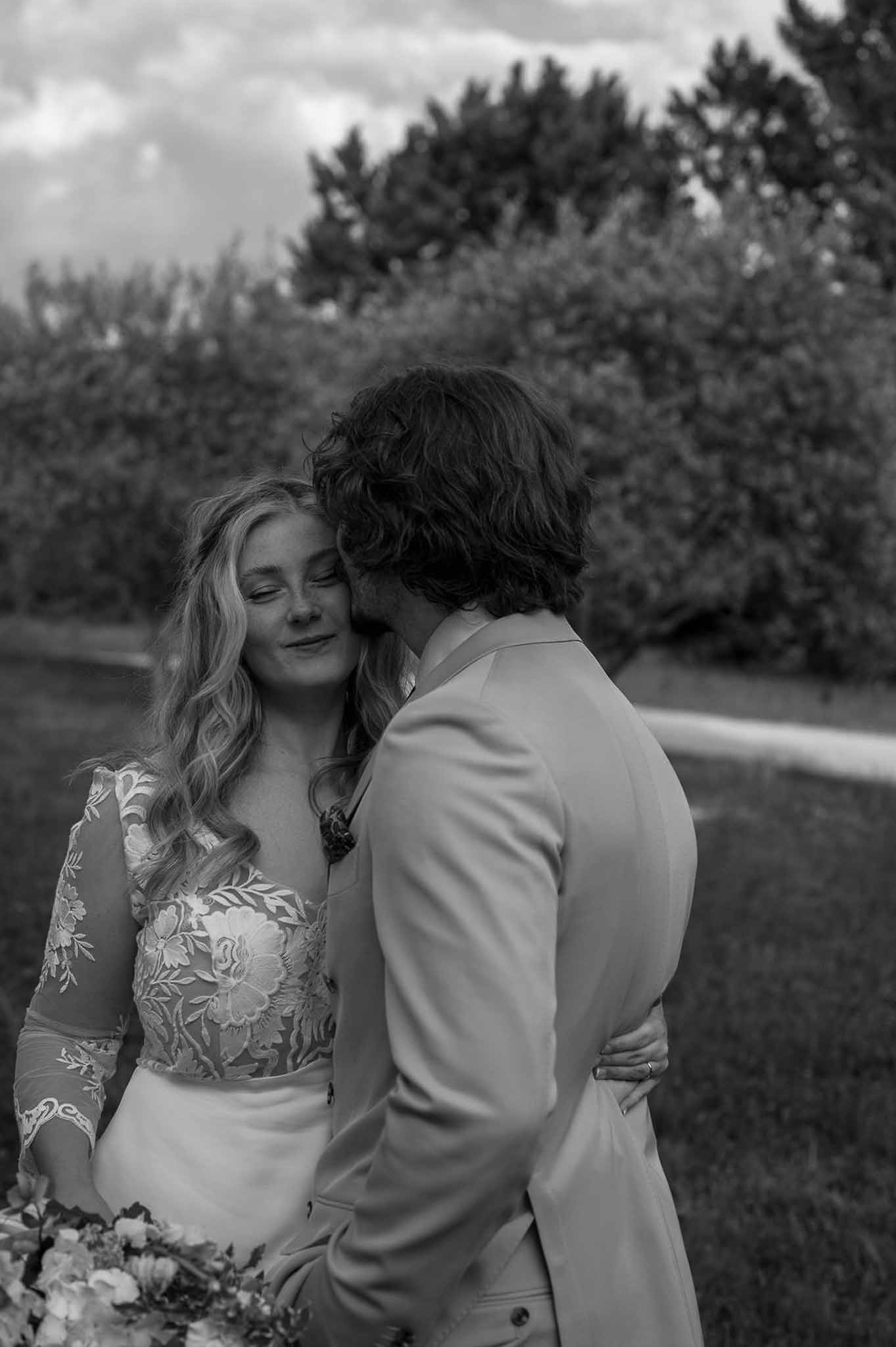 Groom kissing bride's temple as she holds mixed bouquet in sheer floral-embroidered gown in B&W