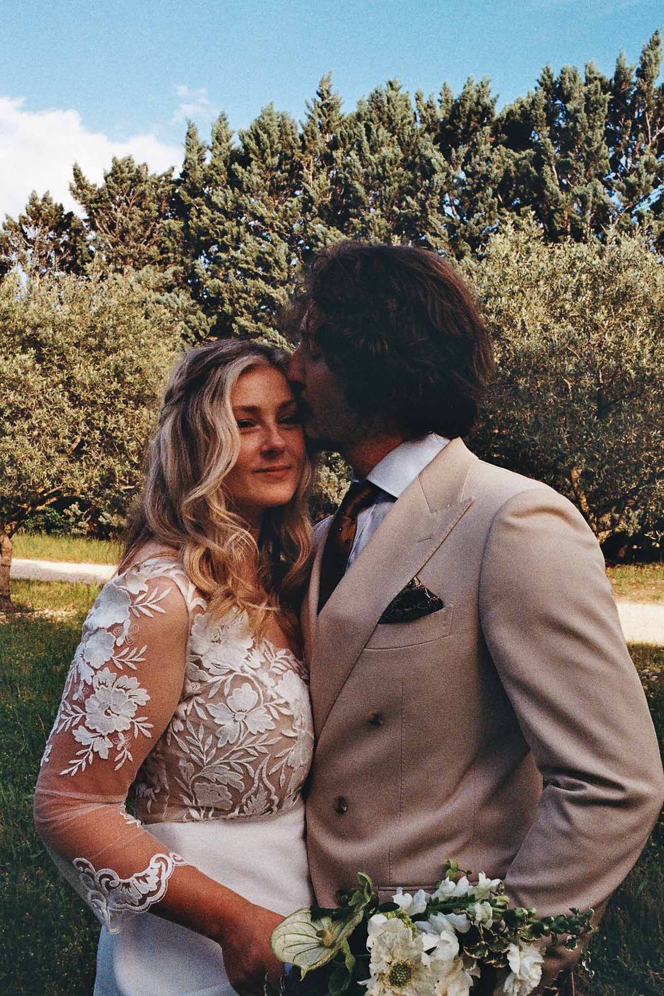 Groom in sand suit kissing bride on temple among olive trees as she holds white flower bouquet