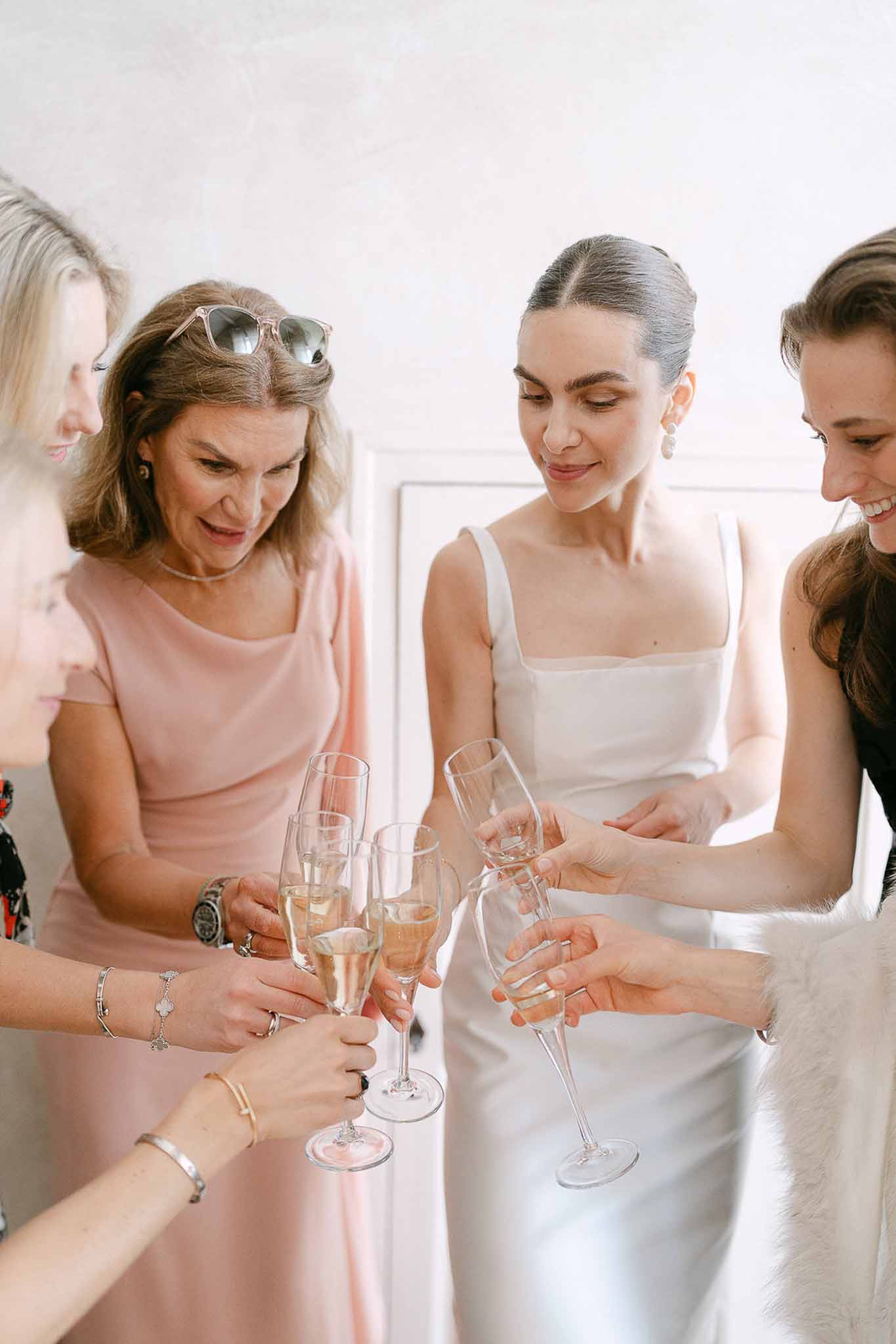Bride and four women clinking champagne flutes during getting ready moment against white wall