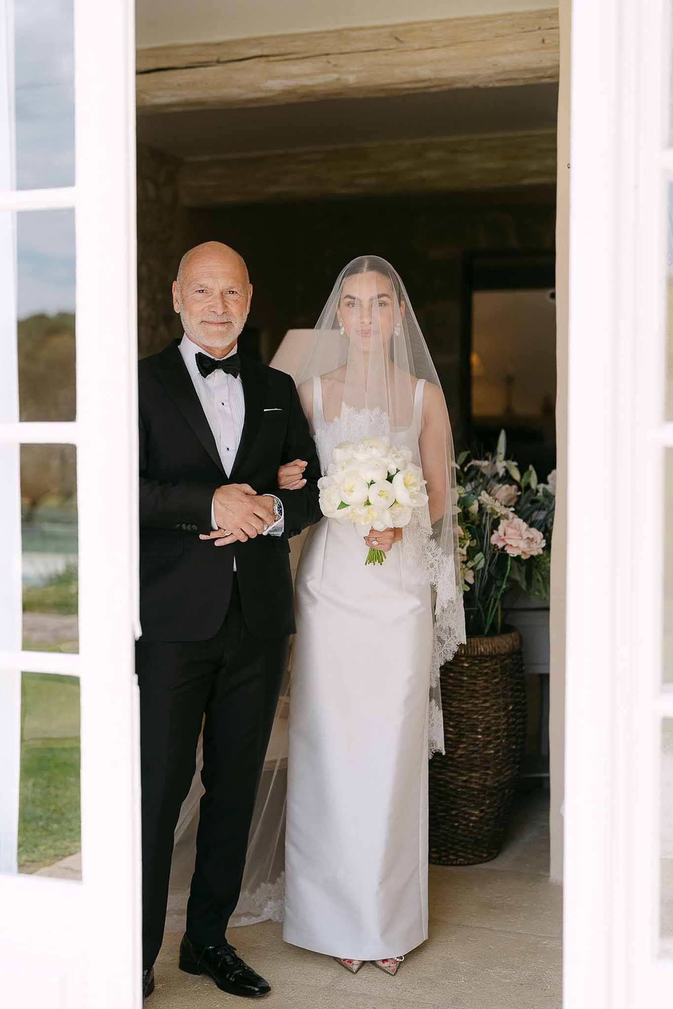 Bride in ivory column gown with lace-edged veil and peony bouquet beside father in chateau doorway