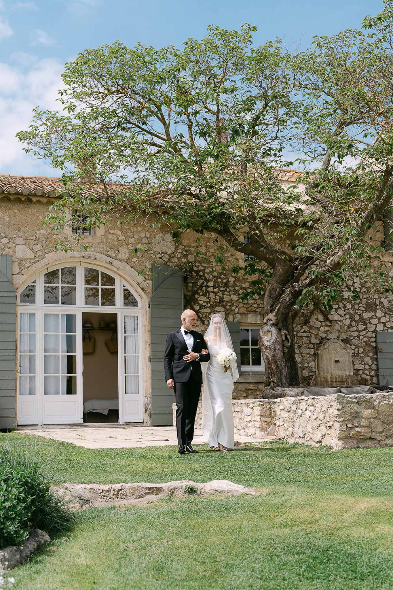 Bride in ivory column gown with cathedral veil and father in tuxedo before Provencal stone farmhouse with blue shutters