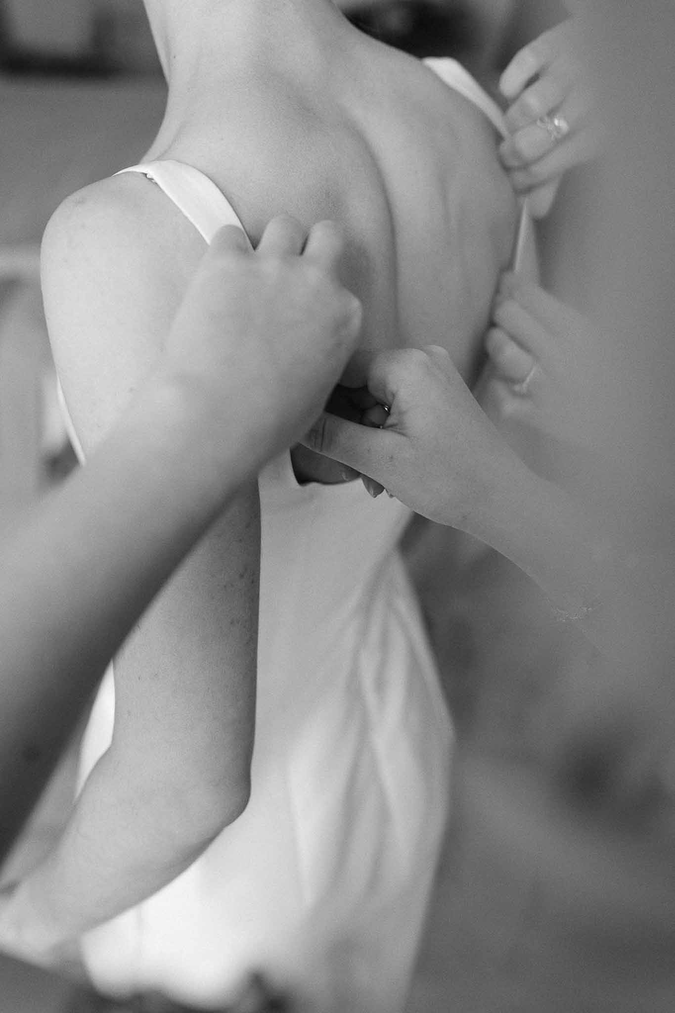 Black and white close-up of hands fastening back of wedding dress during getting ready