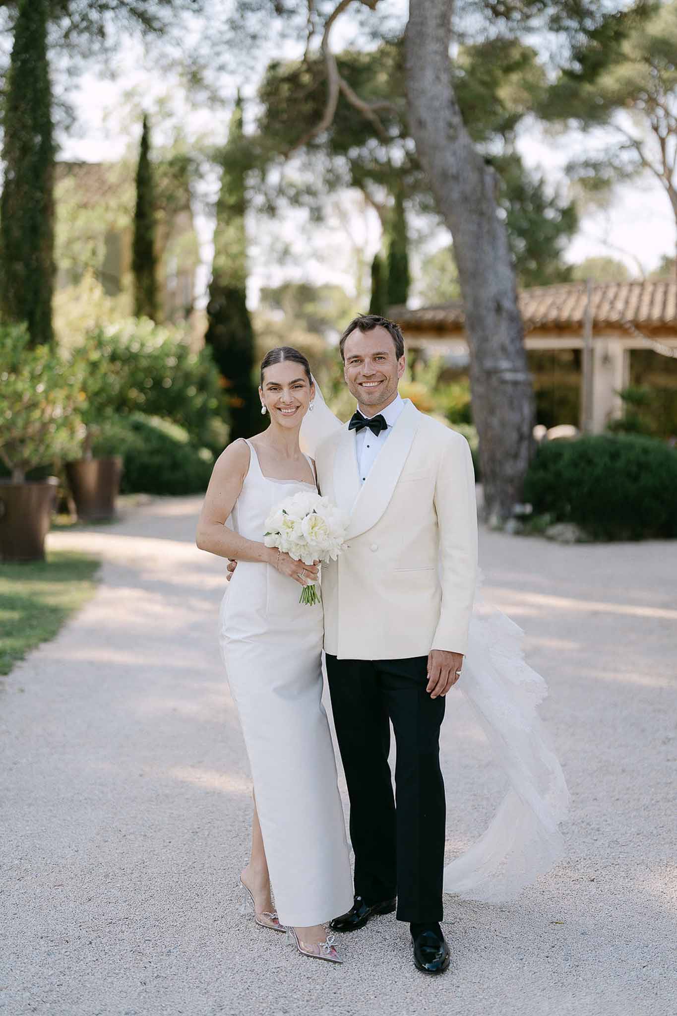 Full-length portrait of bride in ivory column gown and groom in cream dinner jacket on Provencal estate driveway