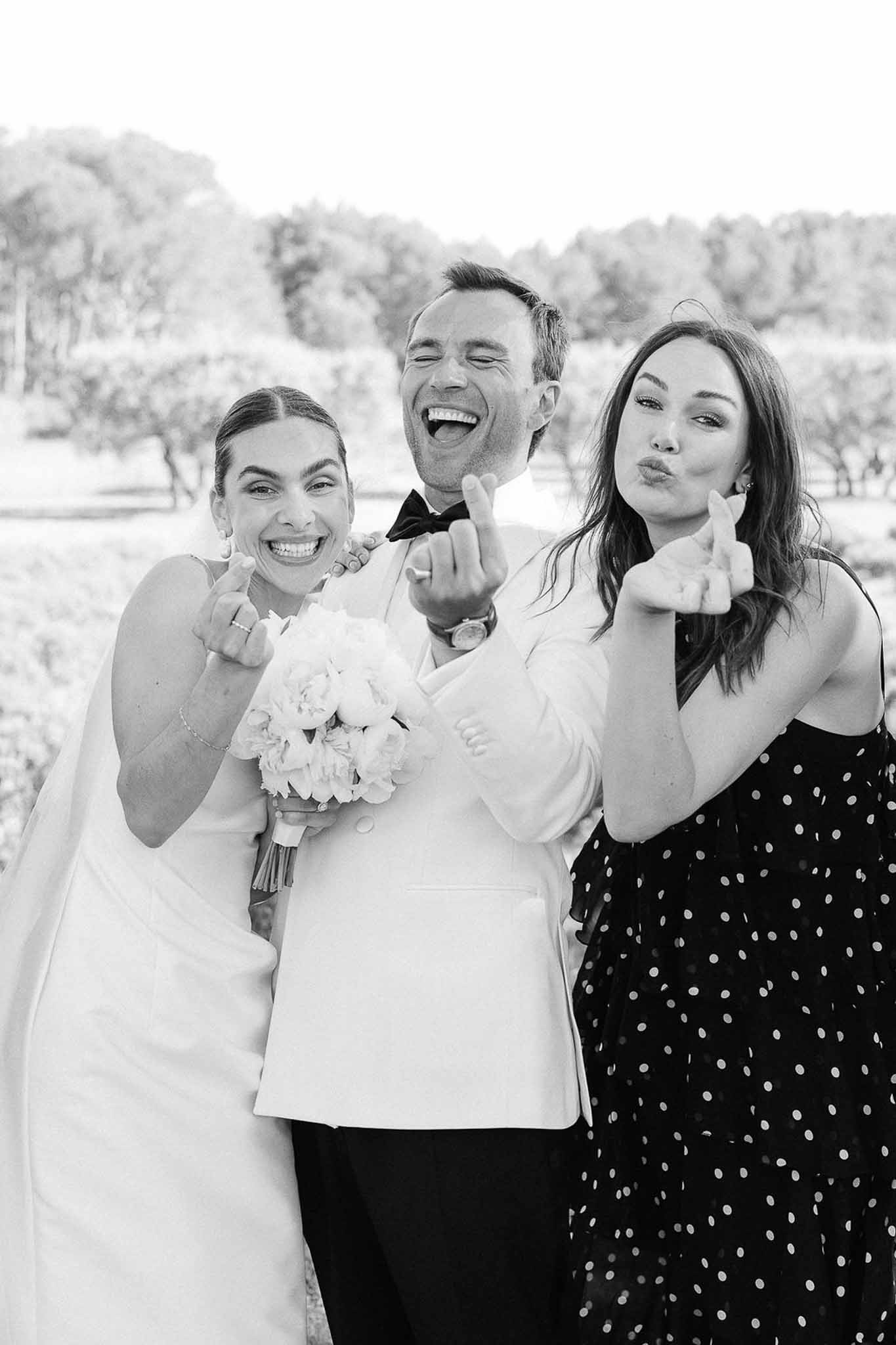 Bride, groom in white dinner jacket, and guest laughing while showing ring fingers in black and white photo