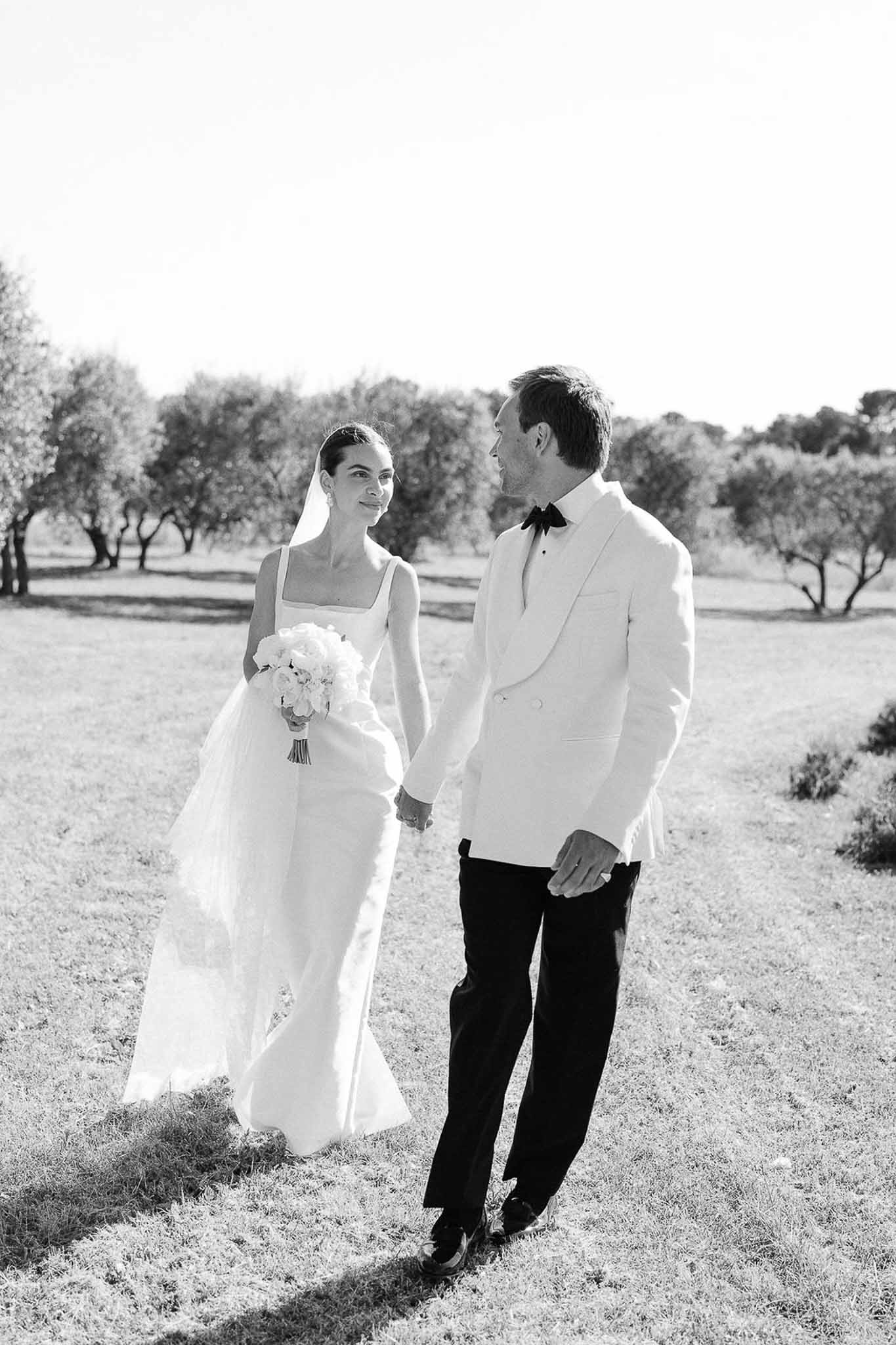 Black and white portrait of bride and groom walking hand in hand through tree-lined outdoor setting