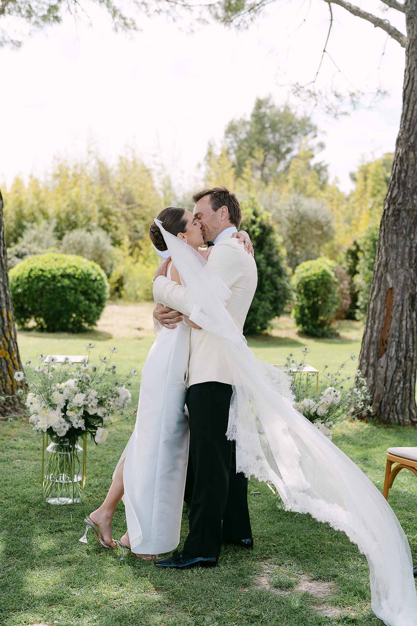 First kiss with sweeping veil bride in ivory column gown groom in ivory jacket with dahlia aisle arrangements