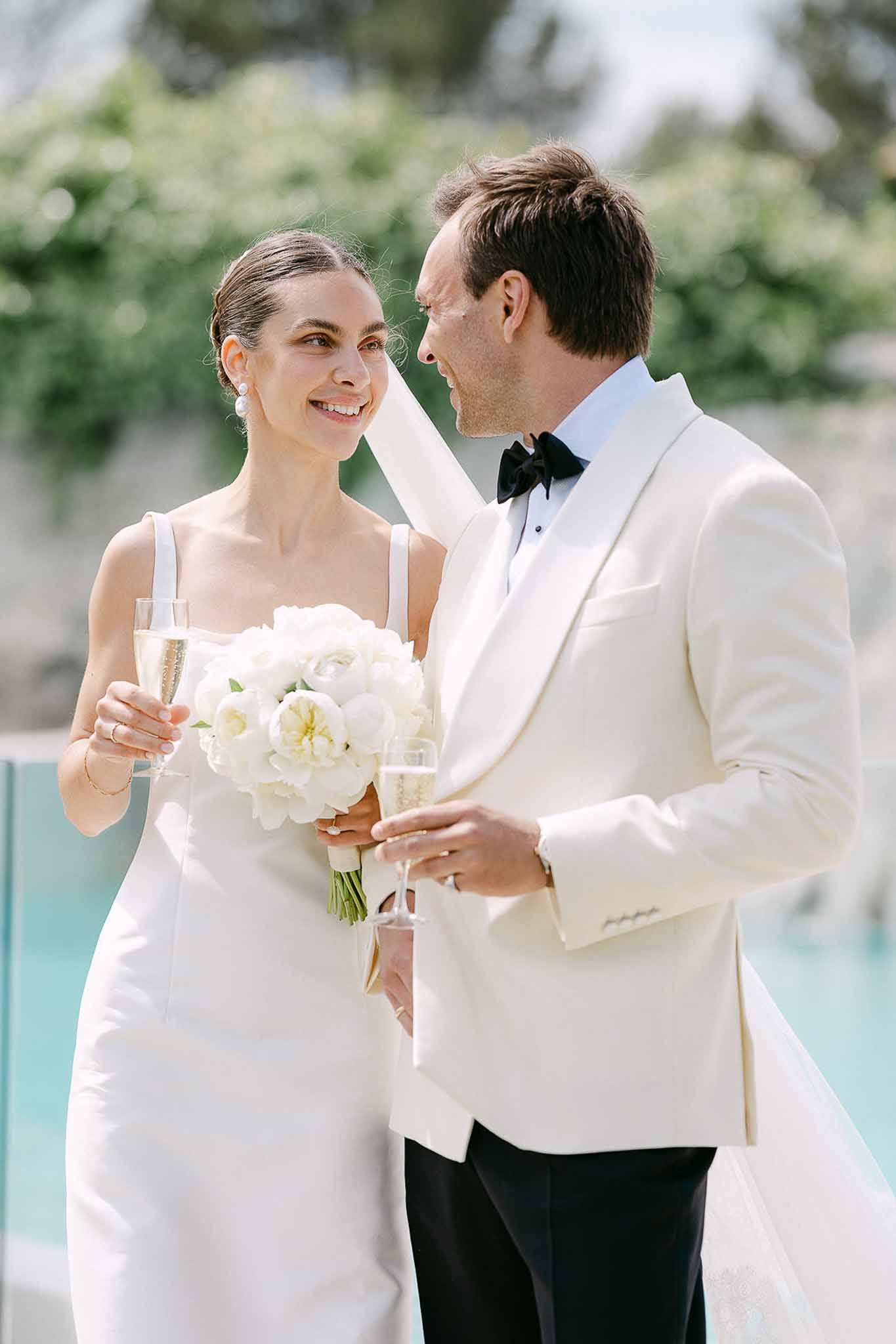 Bride in ivory slip gown and groom in ivory dinner jacket smiling at each other holding champagne by pool