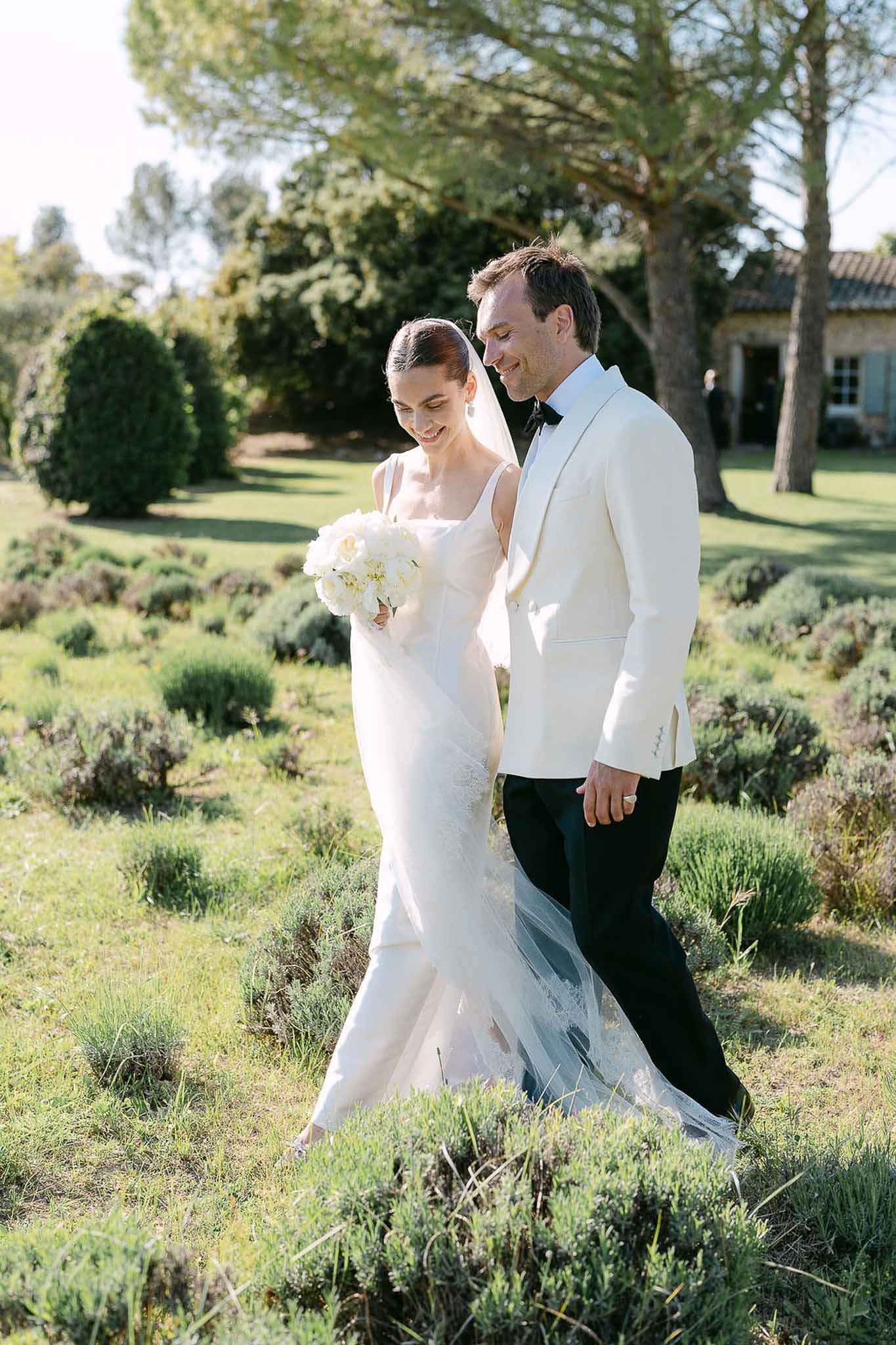 Bride and groom walking through lavender garden at French country estate, bride in ivory slip gown with white peony bouquet