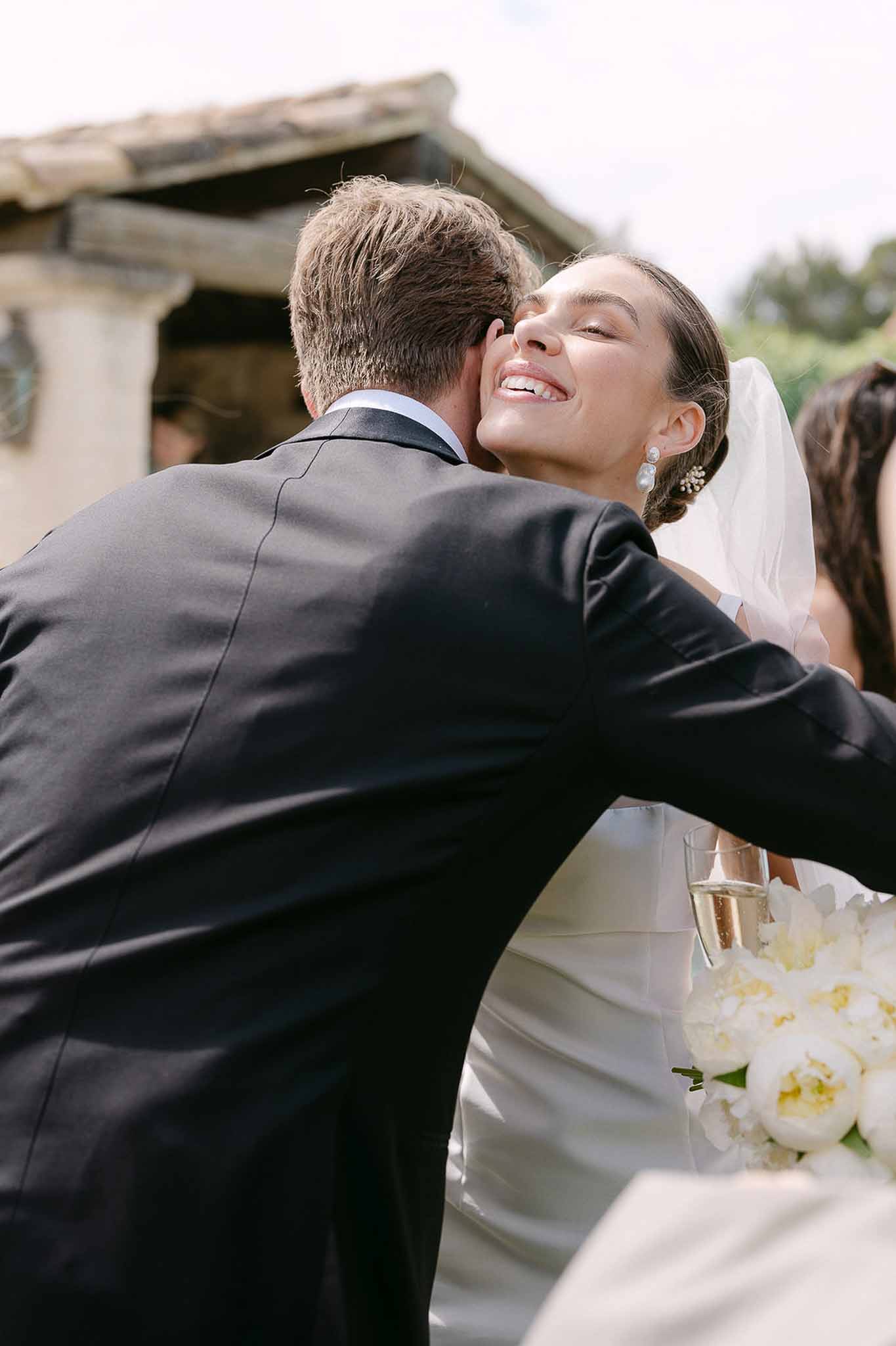 Bride with champagne and white peony bouquet embracing guest outside rustic stone building