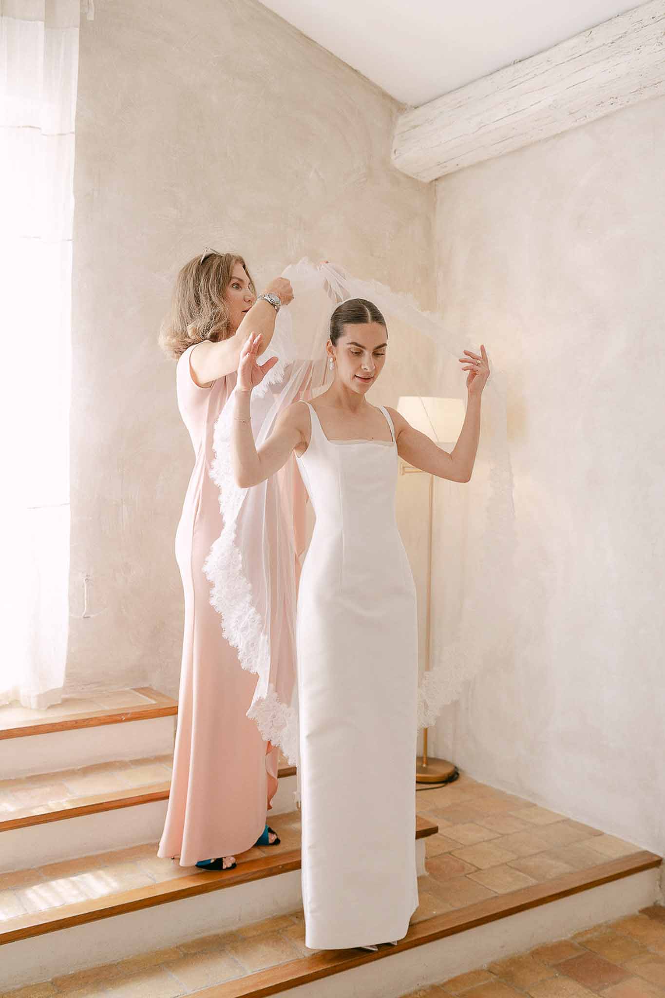 Woman in blush pink dress placing lace-trimmed tulle veil on bride in white satin column dress indoors