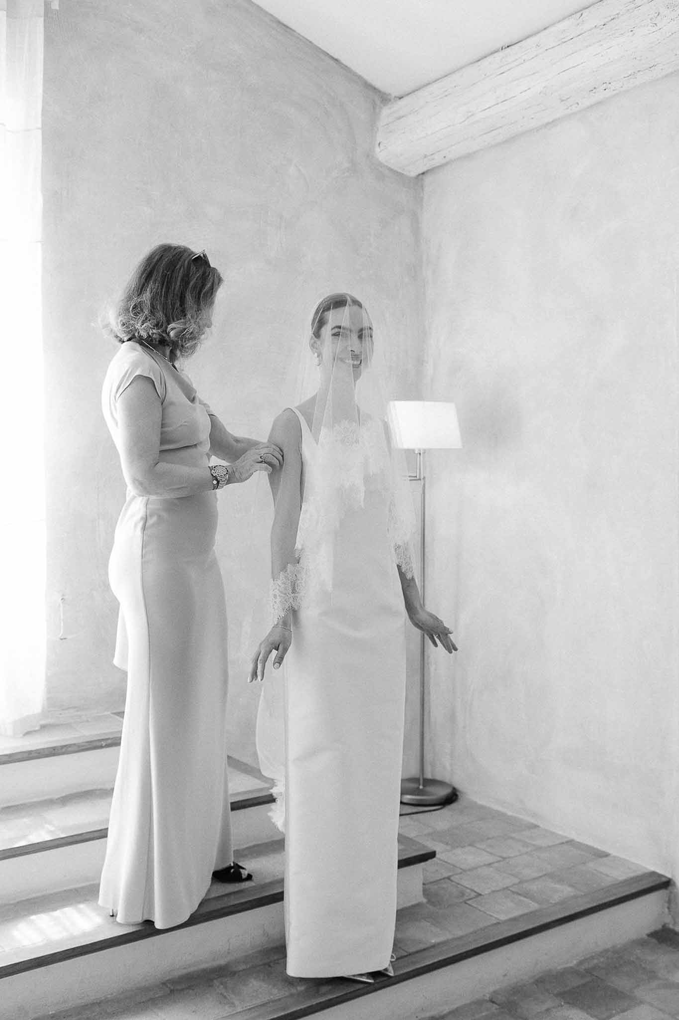 Mother adjusts bride's lace-trimmed veil in bright Provencal room with plaster walls in B&W
