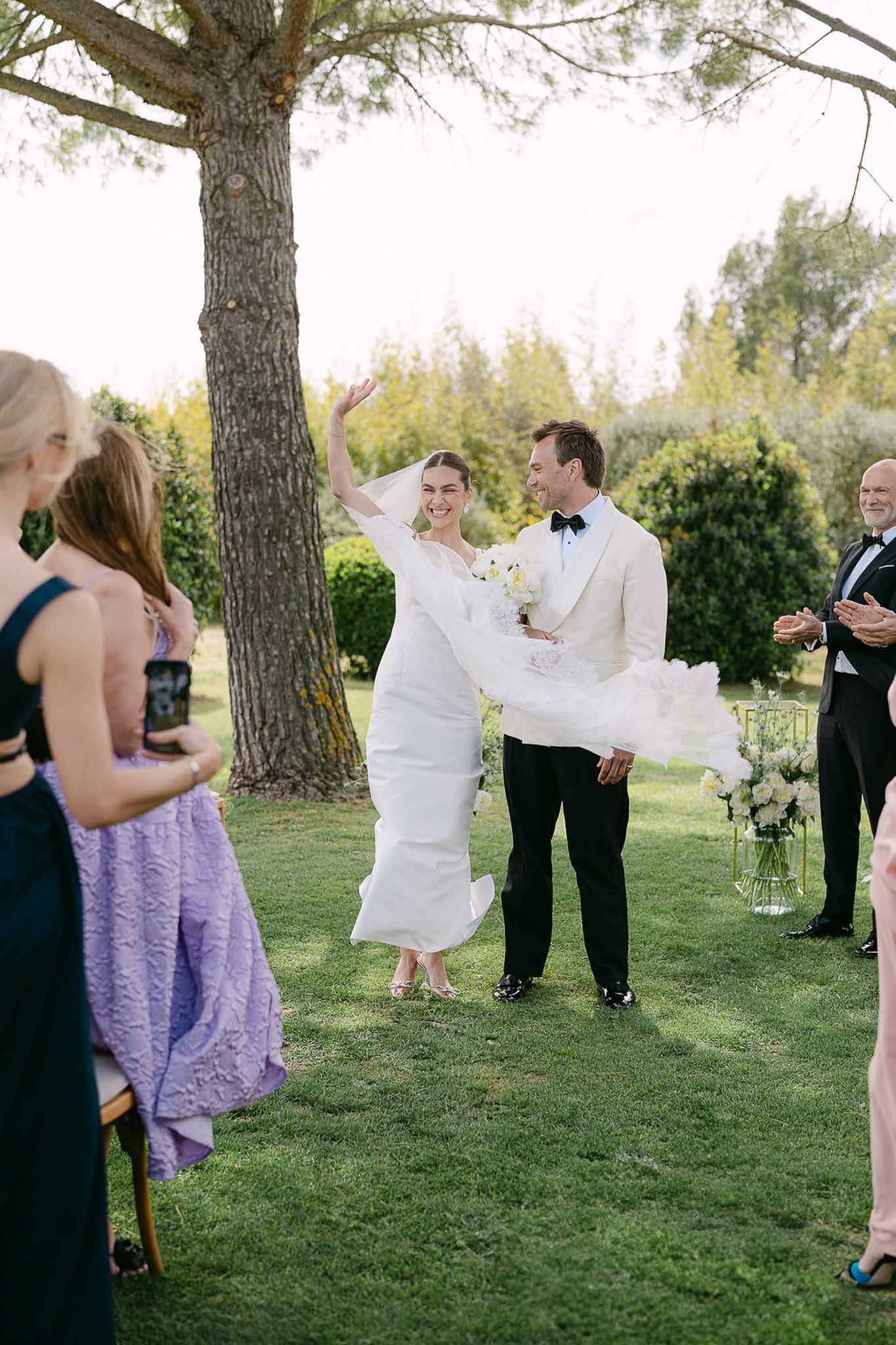 Bride raising arm in celebration during recessional as guests clap on outdoor lawn