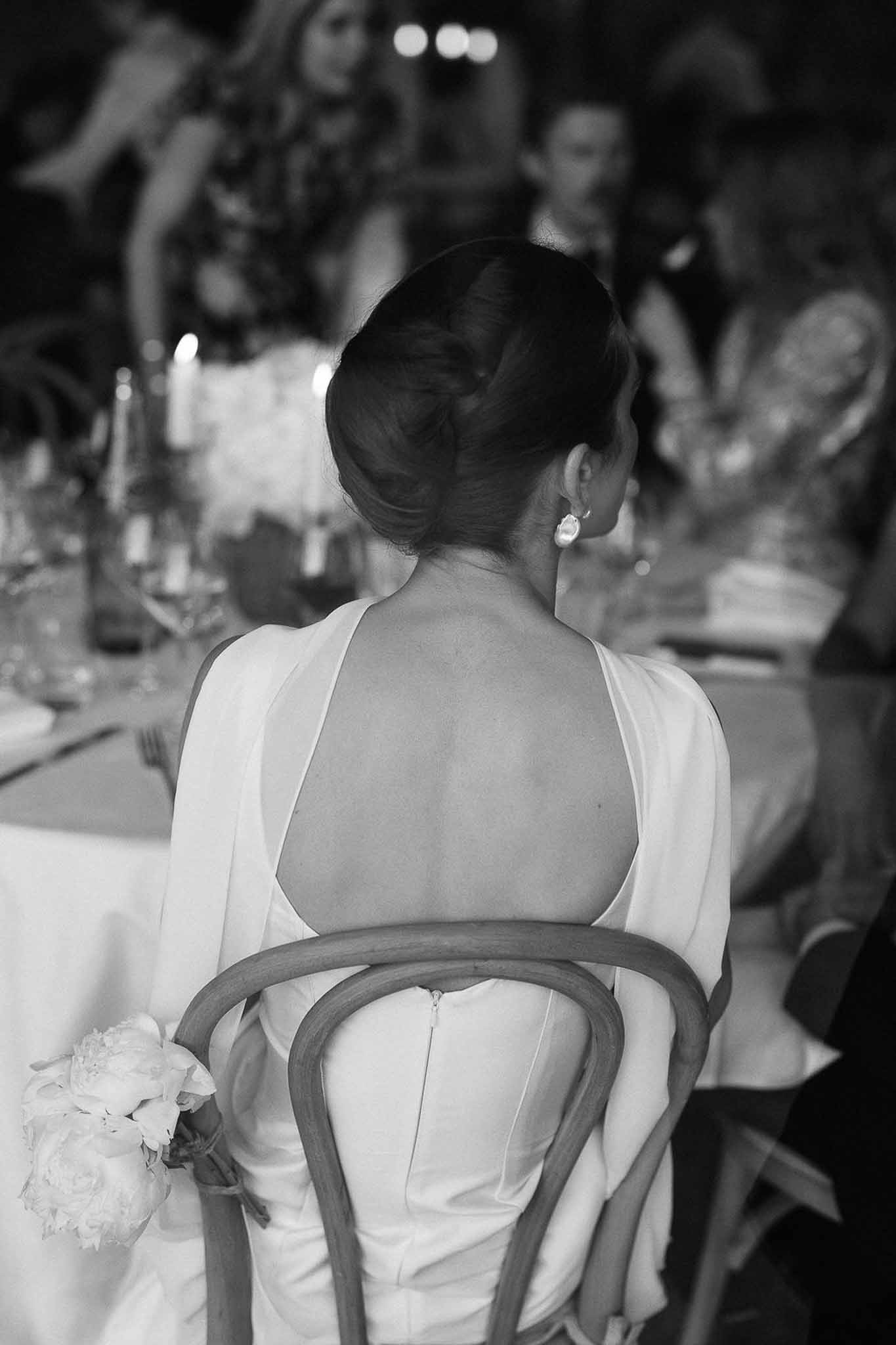 Black and white portrait of bride from behind in open-back gown with cape sleeves at candlelit reception table