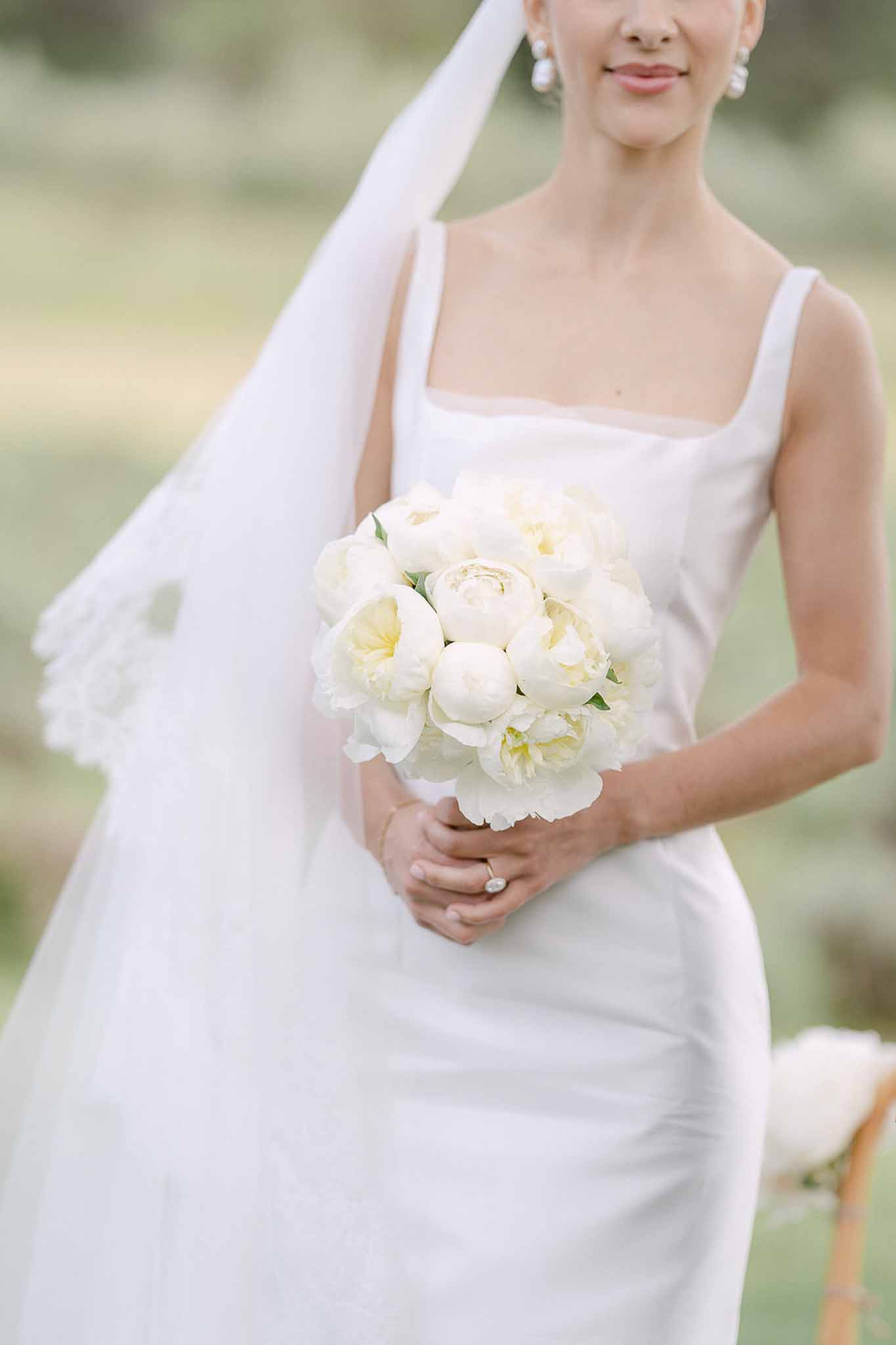 Close-up of bride holding cream and white peony bouquet wearing square-neck gown with lace-edge veil