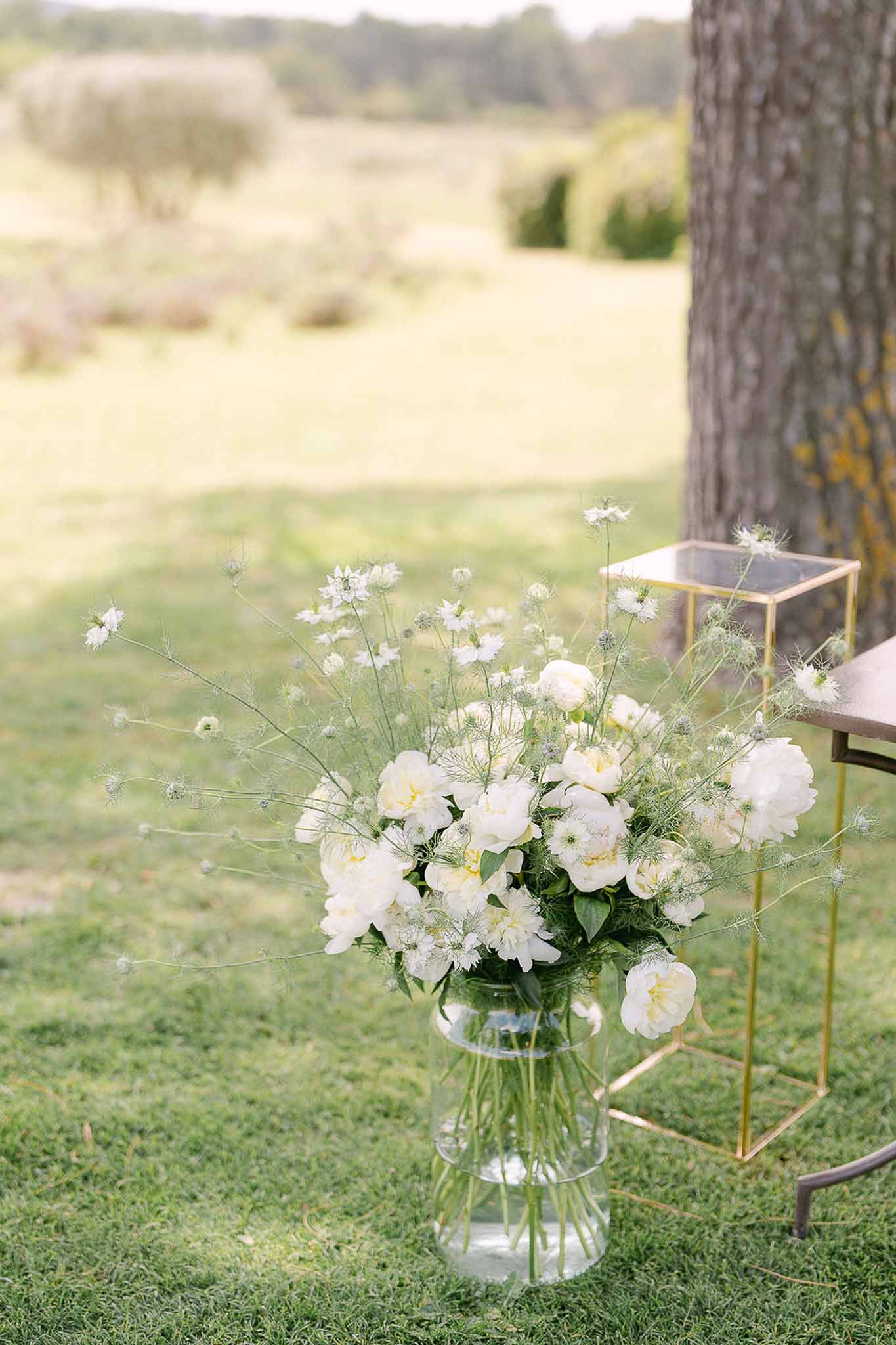White peonies and cosmos in glass vase beside gold side table at outdoor ceremony