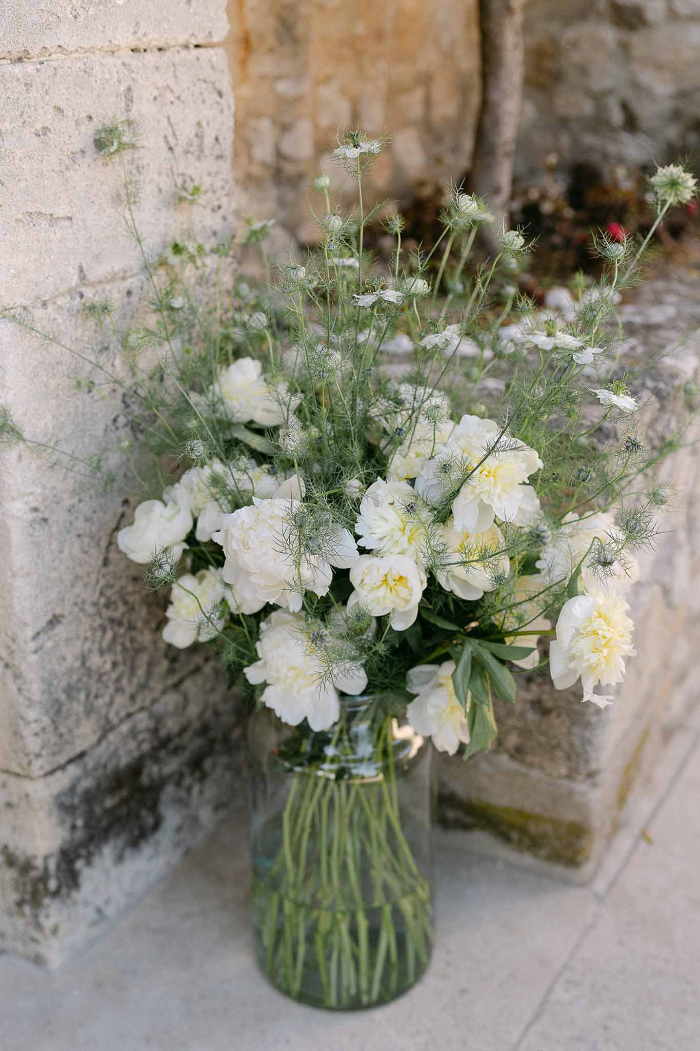 Cream peonies and white nigella in clear mason jar on weathered stone surface against stone wall