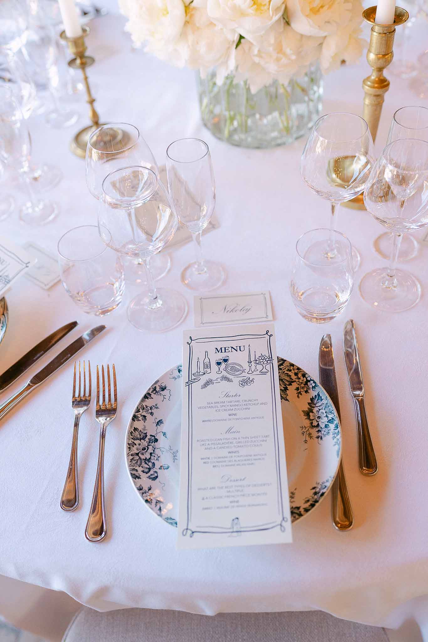 Place setting with blue and white toile charger plate, illustrated menu card, brass candlesticks, and cream peonies