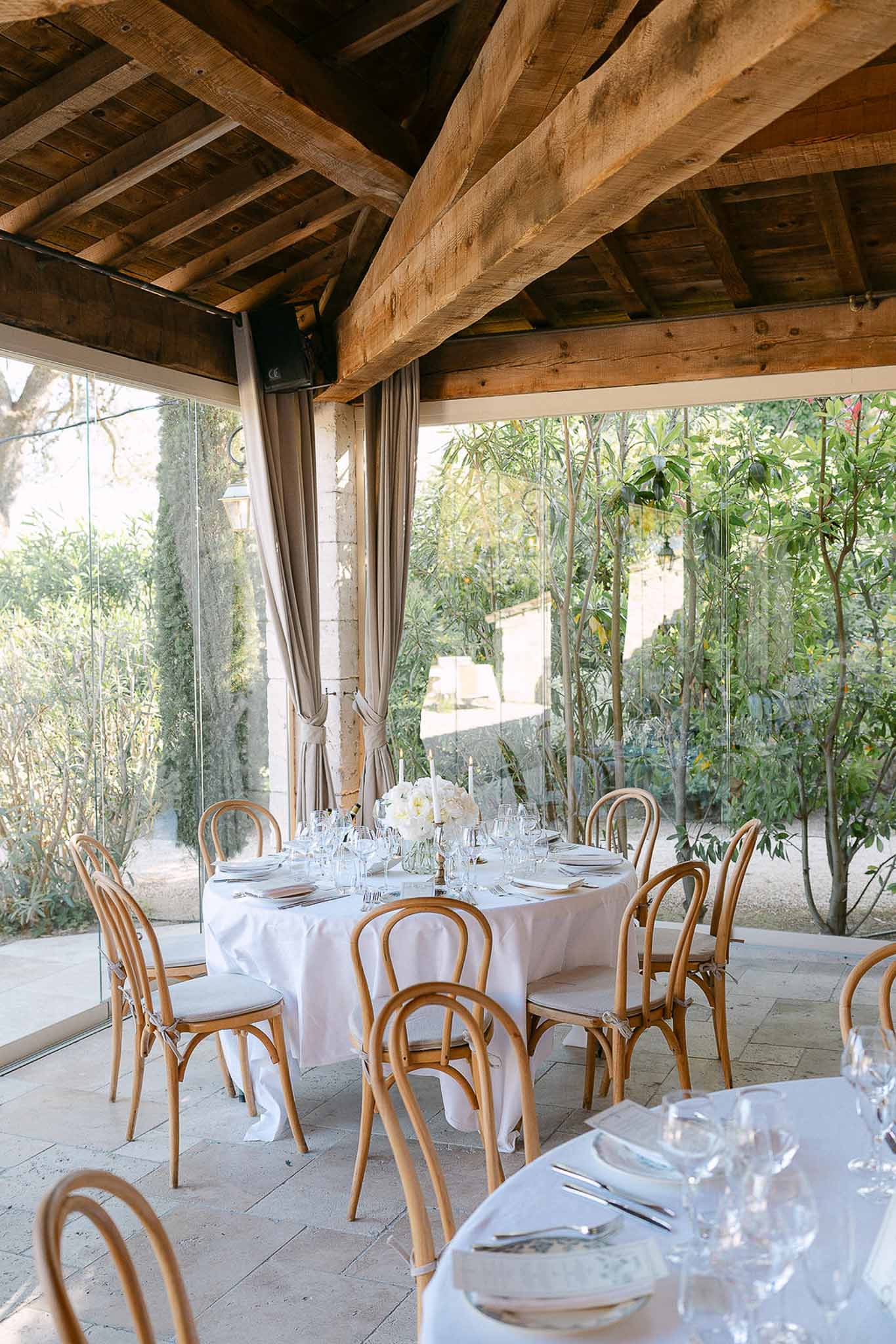 Glass pavilion wedding reception with round tables, bentwood chairs, white floral centerpieces and taper candles