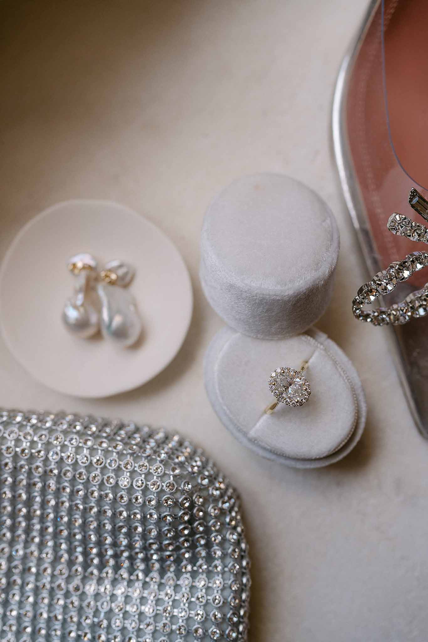 Pear-cut diamond ring in grey velvet box with baroque pearl earrings and crystal clutch on cream surface