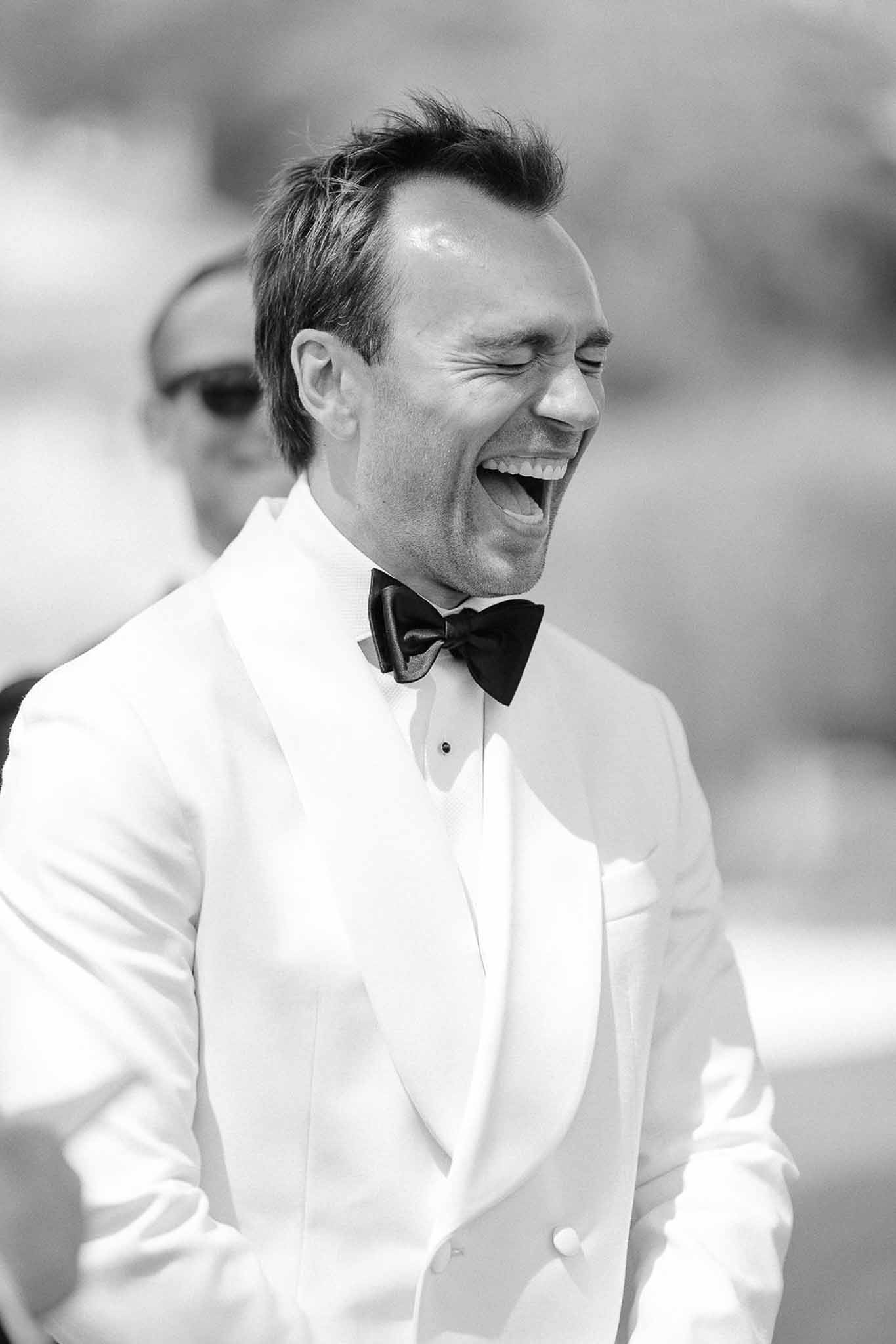 Black-and-white close-up of groom laughing in white double-breasted tuxedo and black bow tie