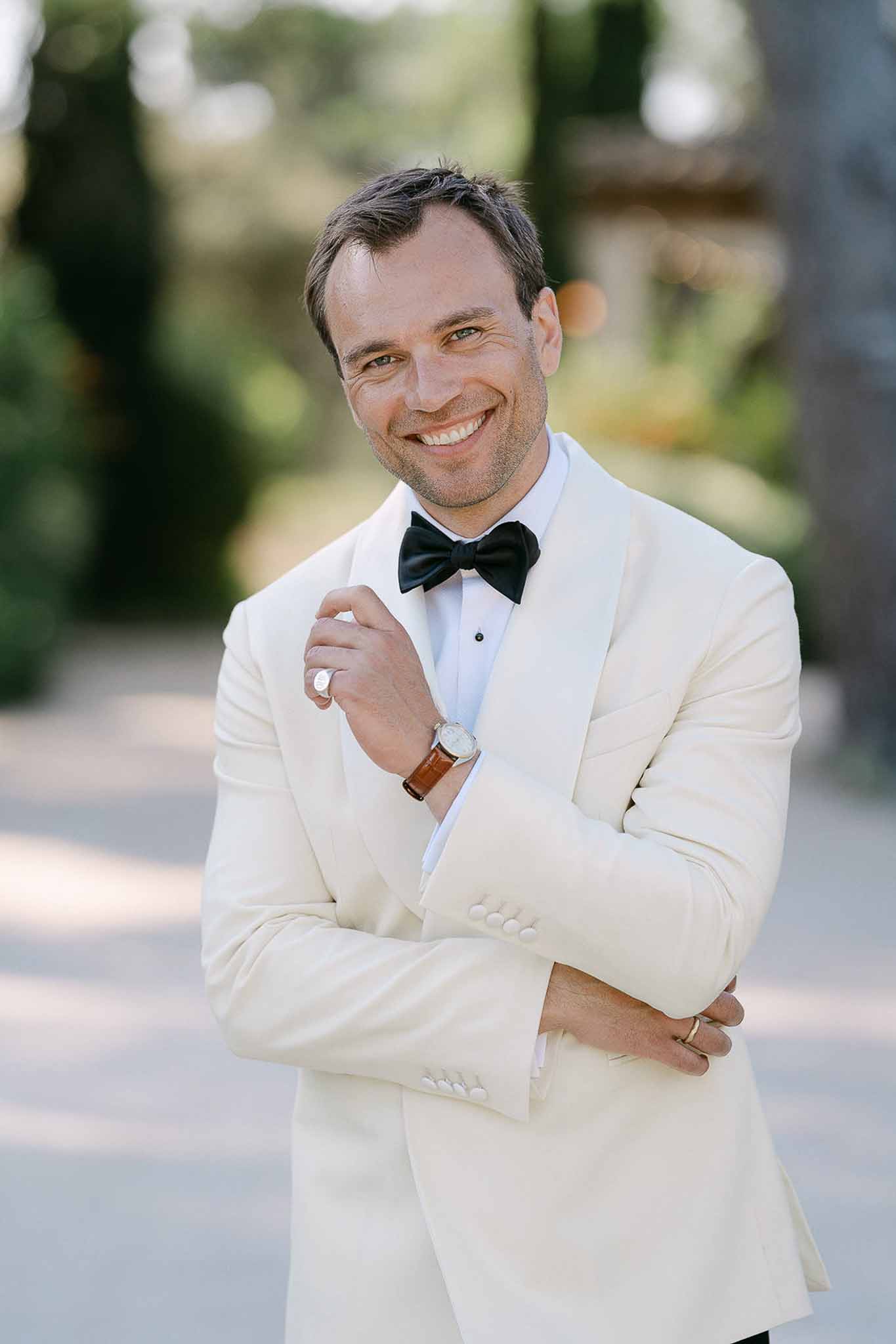 Groom smiling with arms crossed in ivory shawl-lapel dinner jacket on estate grounds
