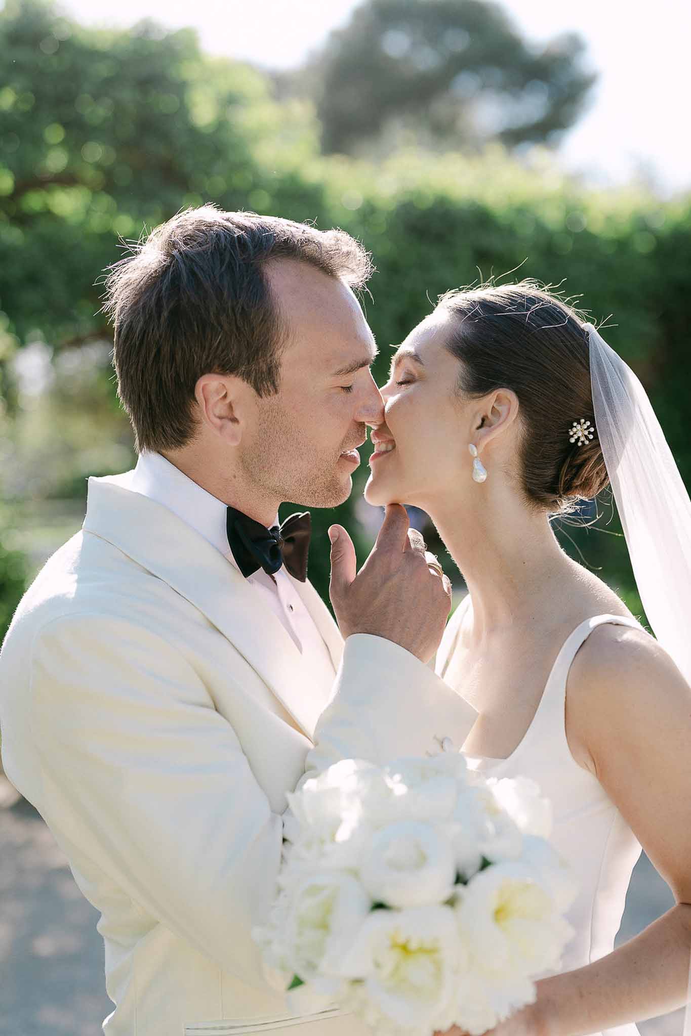 Close-up couple about to kiss bride in ivory gown with cathedral veil and white peony bouquet