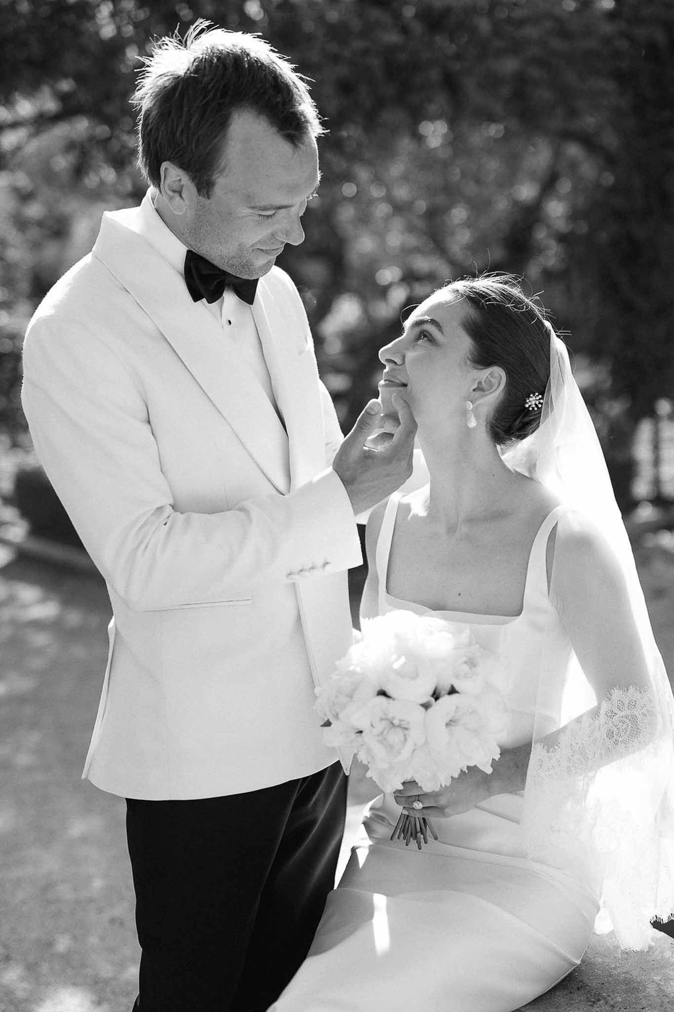 Black and white portrait of groom touching bride's face as she holds peony bouquet in garden setting