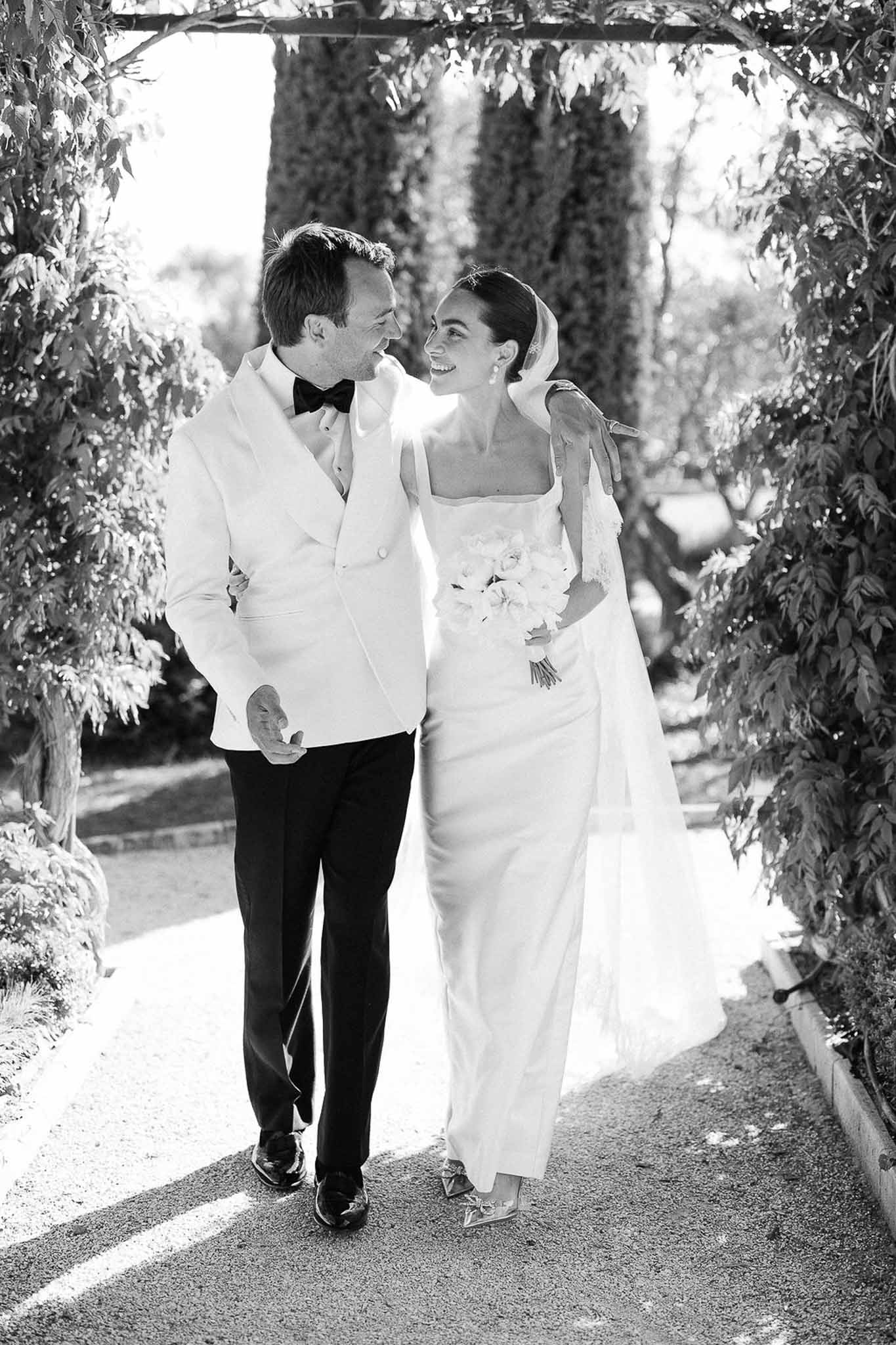 Black and white couple walking arm in arm under leafy pergola with flowing veil and white dinner jacket