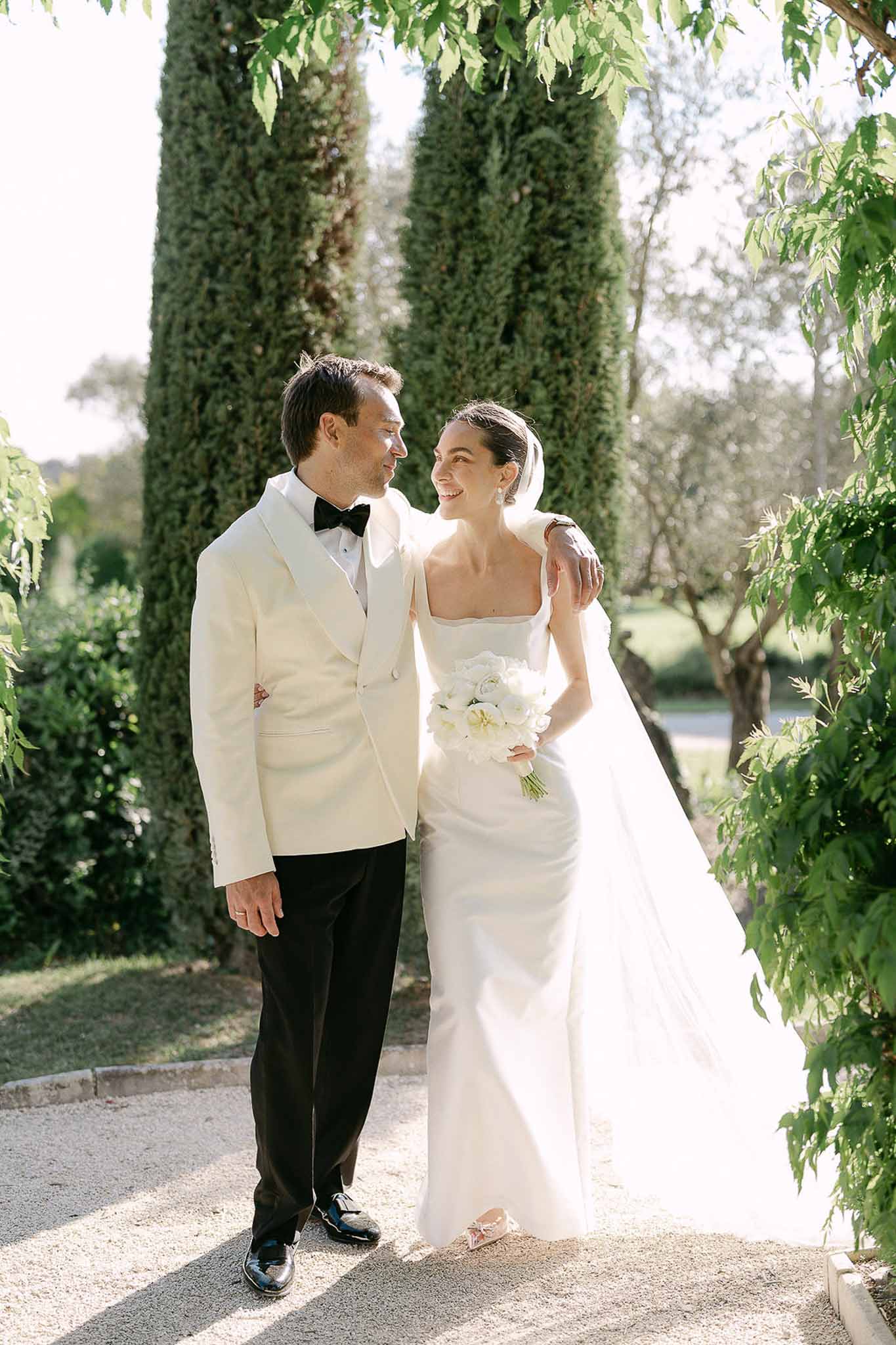 Couple on cypress-lined gravel path with groom in ivory dinner jacket and bride holding peony bouquet