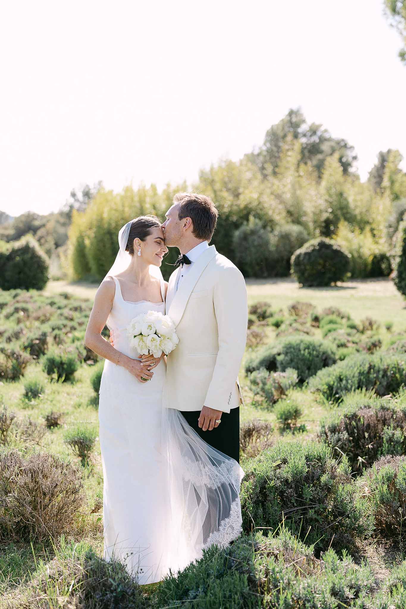 Groom kissing bride's forehead as she holds white peony bouquet in herb garden