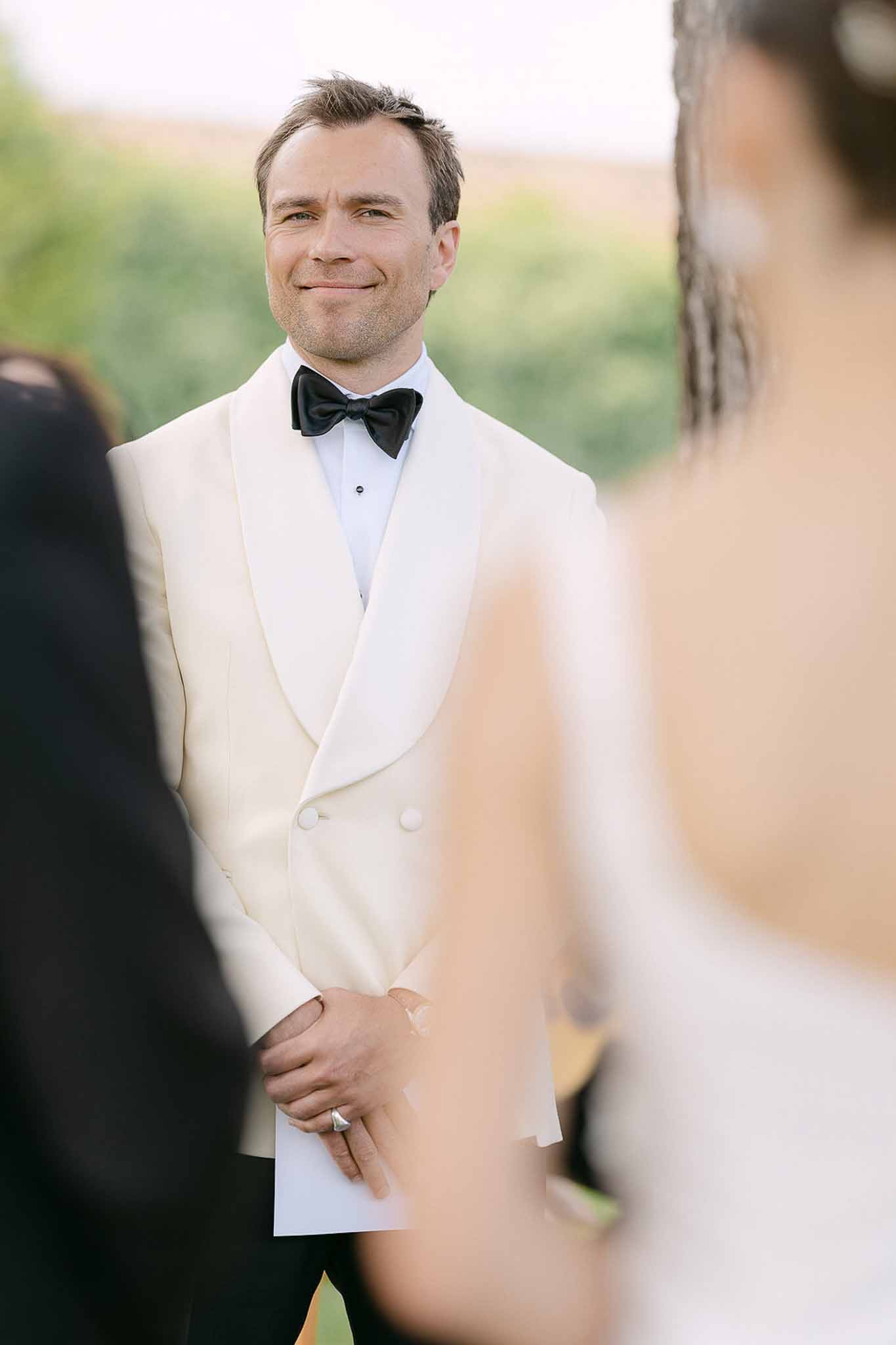 Groom in an ivory dinner jacket watching the bride approach during an outdoor countryside ceremony holding his vows