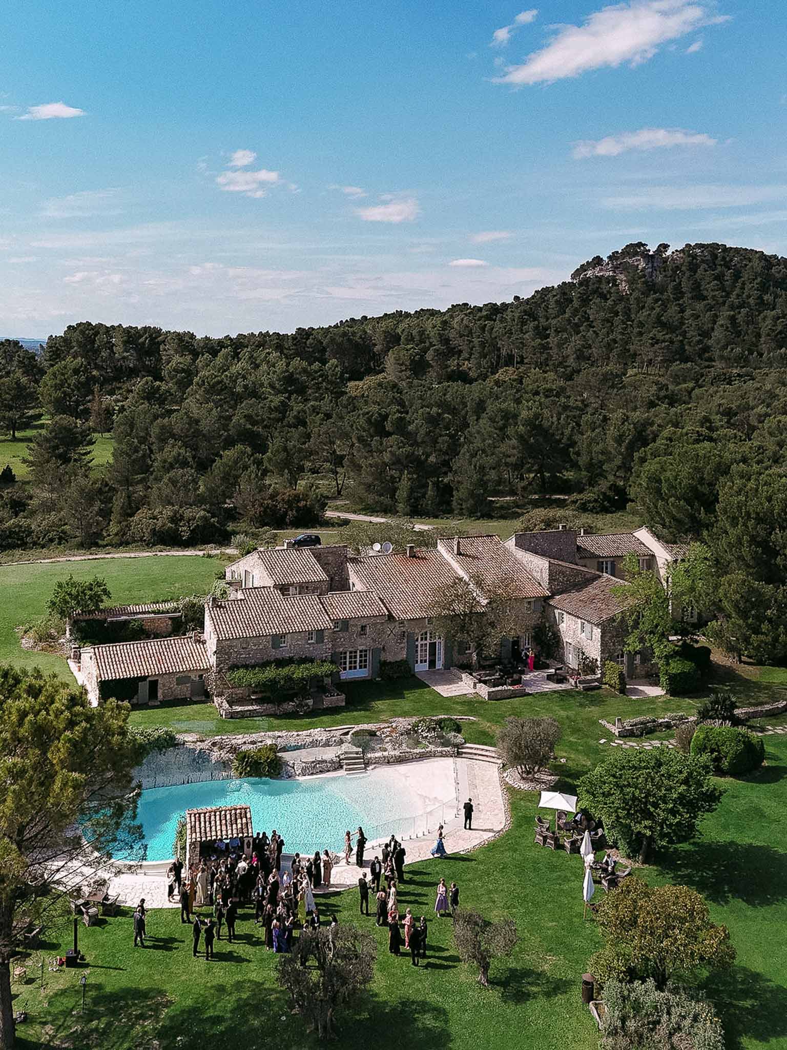 Aerial view of sixty guests at cocktail hour around freeform pool at Provencal mas with wooded hills