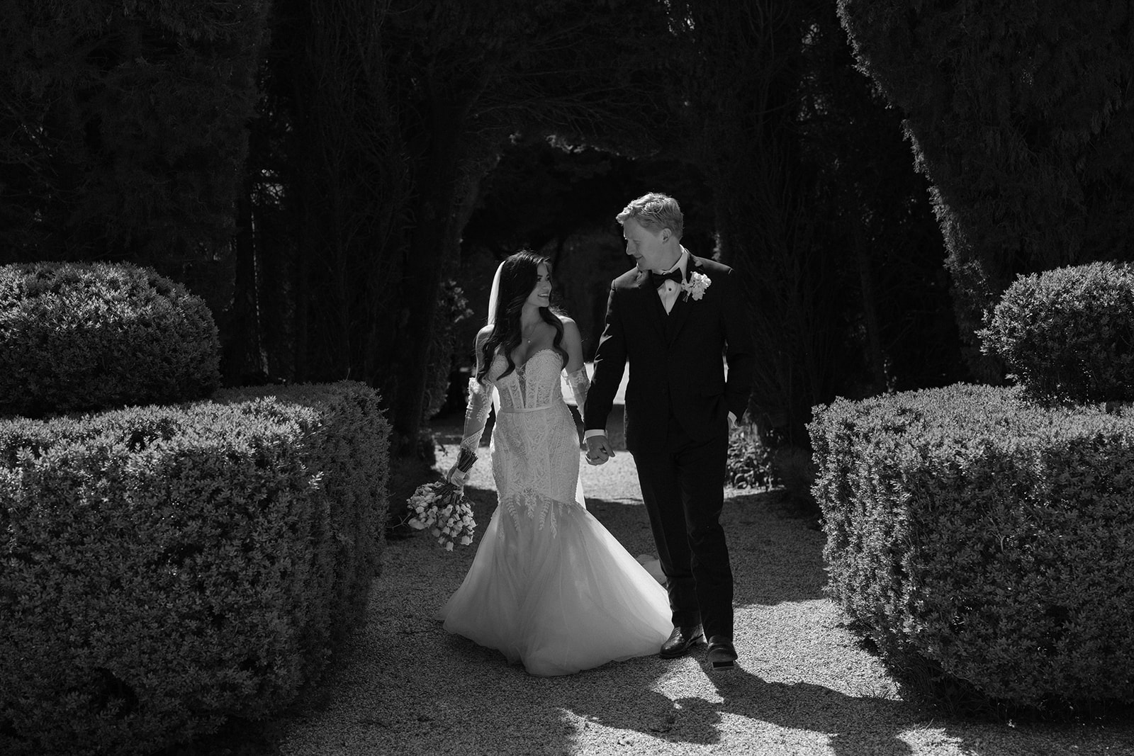 Black and white portrait of couple walking hand-in-hand along gravel path between cypress trees